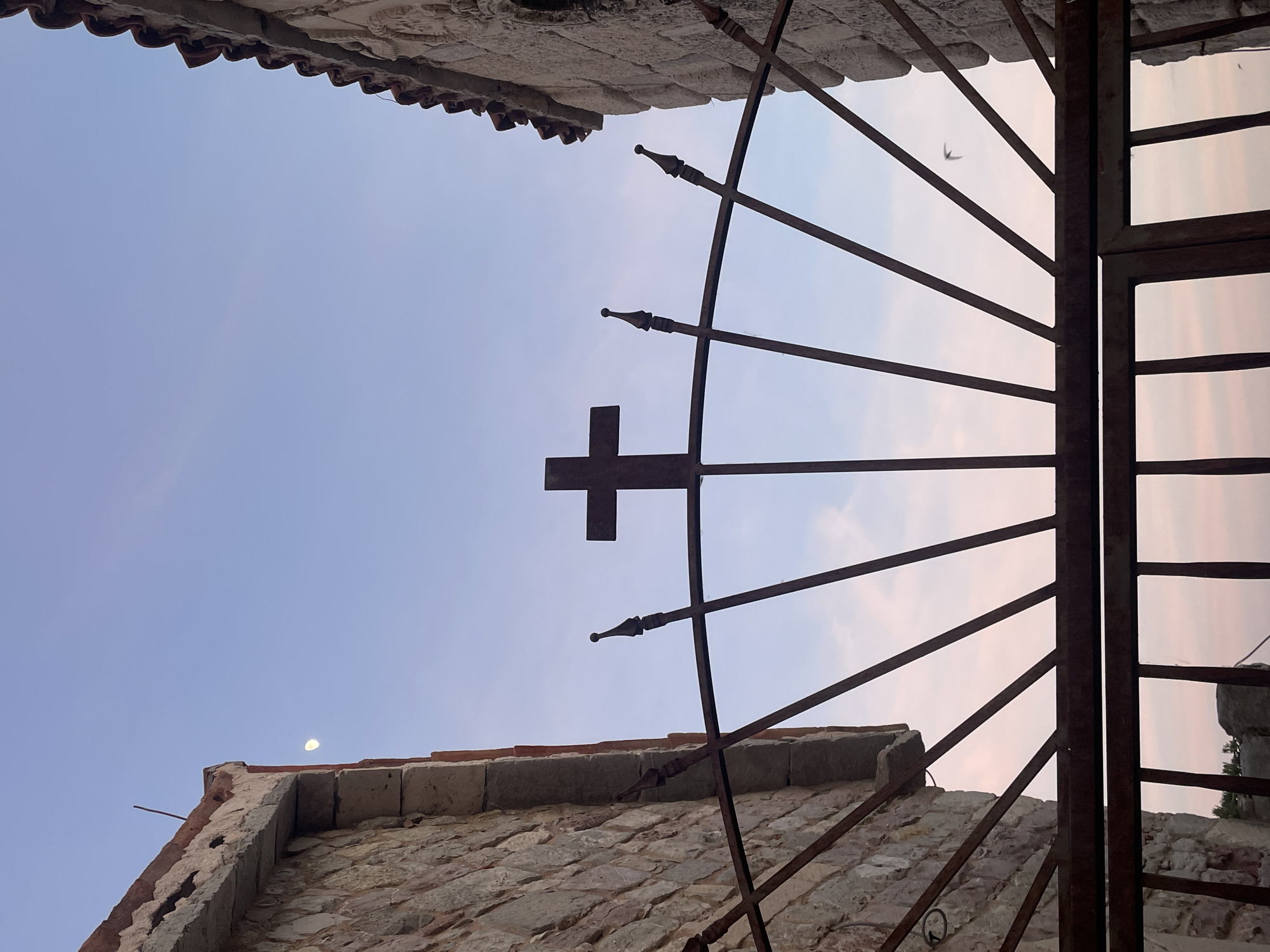The photograph depicts an upward view through a wrought iron gate adorned with a cross, framed by stone buildings. Above, a light blue sky is visible. A bird is captured in flight between the bars of the gate. The buildings appear to be constructed of aged stone and brick, with red tile roofing. A sliver of the moon can be seen. The location is not specified.