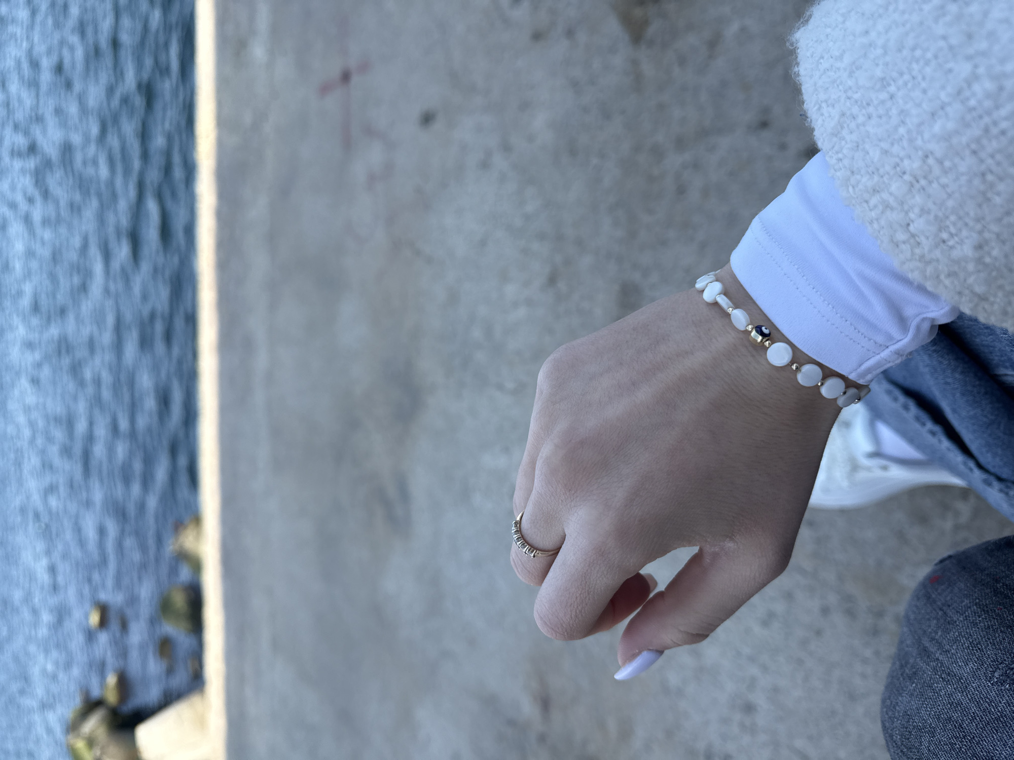 A close-up shot shows a person's hand and wrist adorned with jewelry, positioned against a backdrop of a concrete structure and a body of water.
The hand wears a gold ring on the ring finger and a bracelet consisting of white and gold beads on the wrist. The fingernails are painted a light lavender color. The arm is clothed in a white, long-sleeved shirt, partially obscured by a fuzzy white jacket or sweater. Blue jeans can be seen partially within the frame.
The background includes a grey concrete structure, possibly a retaining wall or a section of a building. To the left, there is a body of water with a dark, rippling surface, possibly the Black Sea. Vegetation is visible at the water's edge.
Given the location context of Varna, Bulgaria, it is plausible that the body of water is the Black Sea and the concrete structure is related to the city's coastal infrastructure.