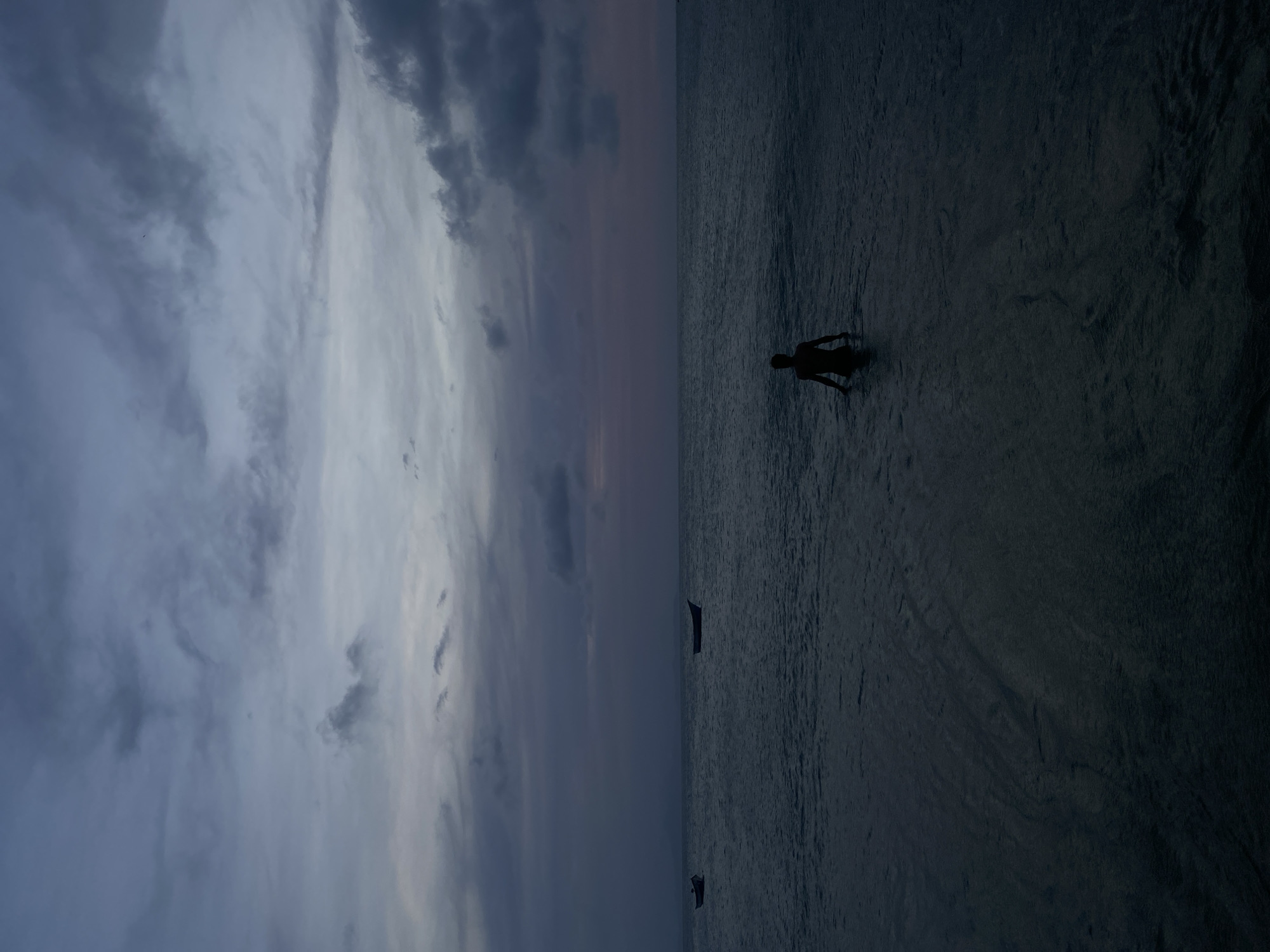 The image depicts a twilight scene in Juan Griego, Venezuela. The sky is filled with layered clouds, ranging in color from gray to purple. A body of water stretches to the horizon. A silhouetted person stands in the water. Two small boats are visible on the water in the distance.