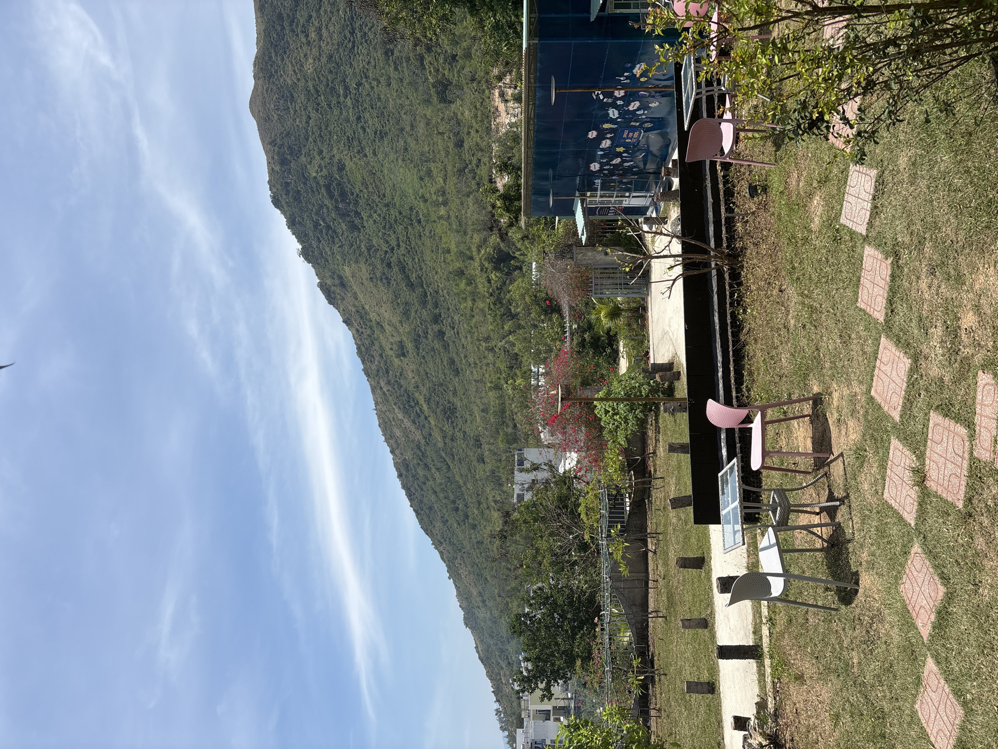 The image depicts a scenic view of a garden and mountain landscape in Nha Trang, Vietnam. In the foreground, there is a well-manicured lawn with pink and white chairs surrounding a glass table. A paved walkway with square tiles cuts through the grass. Beyond the garden, the land slopes upward into a large, green, forested mountain. In the background, white buildings can be seen nestled amongst the trees. A blue building adorned with images of marine life sits beside the garden. The sky is blue with wispy clouds. The overall impression is a peaceful and picturesque outdoor setting.