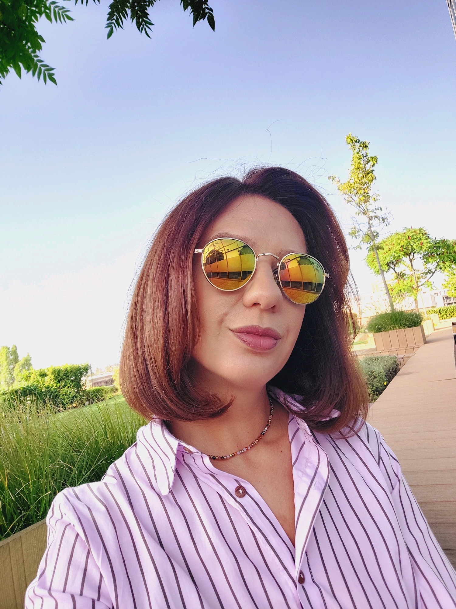 A woman is posing for a selfie outdoors in the city of Sofia, Bulgaria. She has medium-length brown hair, wears round, mirrored sunglasses, and a light pink striped button-up shirt. She is also wearing a beaded necklace. The background shows a clear blue sky, some green foliage and trees, and a wooden walkway.