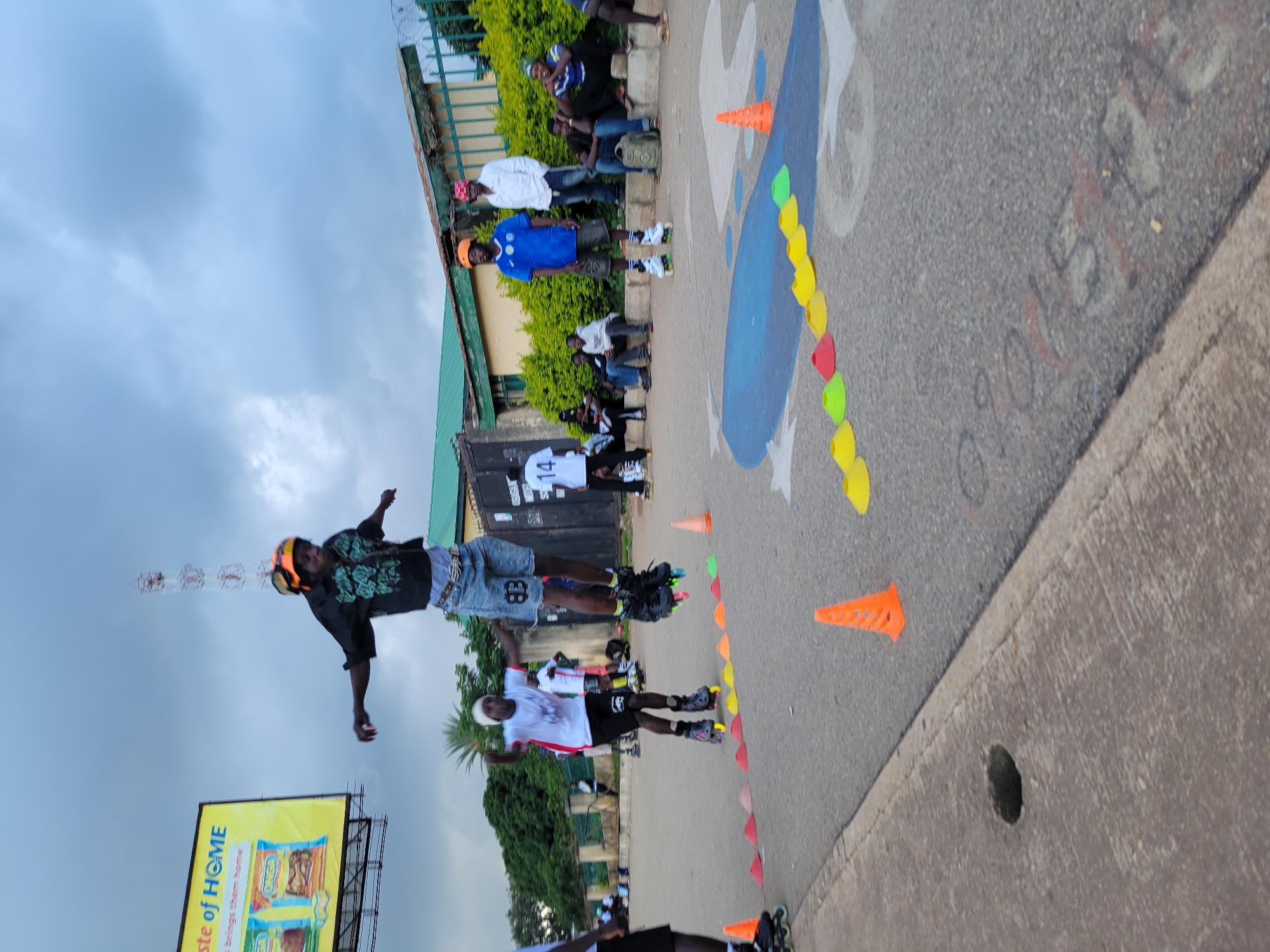 In Jos, Nigeria, a young male rollerblader in mid-air, wearing an orange helmet and a black patterned shirt over denim shorts, performs a jump over a line of multi-colored cones. He is part of a group engaged in rollerblading activities on an asphalt surface marked with blue paint and some graffiti. Several other rollerbladers, some in athletic wear and helmets, are visible in the background. Spectators are seated on a low wall and a raised curb to the right, observing the event. The environment features a green fence, lush bushes, and a building with a green roof, under a cloudy sky. A billboard advertising "Taste of HOME" is visible in the upper left corner of the image.