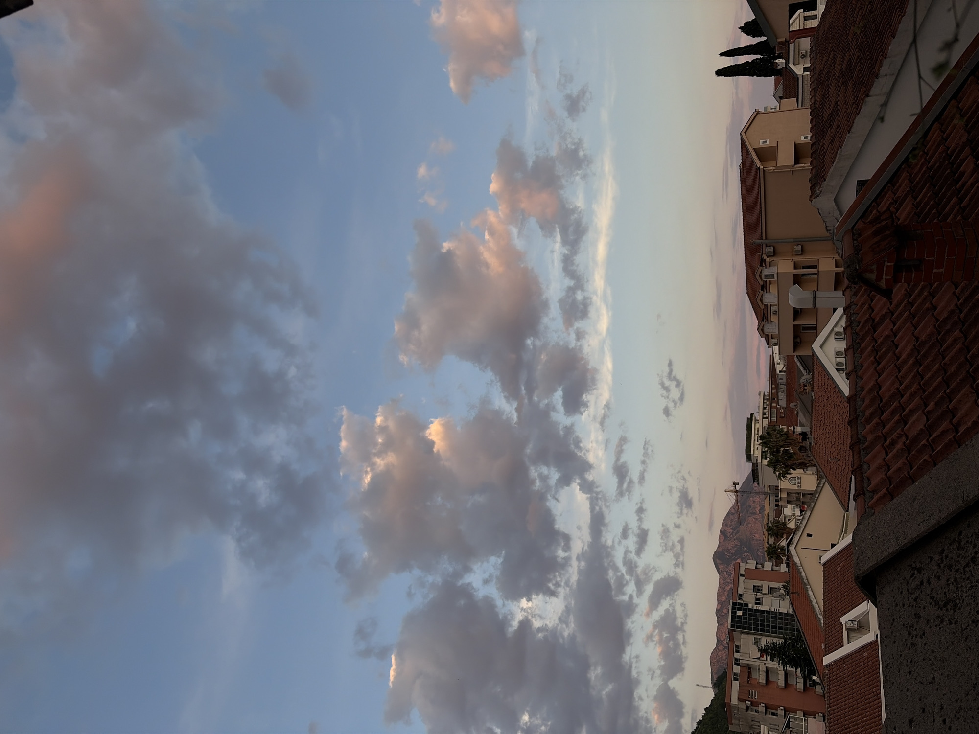 The image shows a view of Budva, Montenegro at dusk. In the foreground, there are rooftops of red-tiled buildings. In the middle distance, there are more buildings, including some with white walls and balconies. In the background, there is a mountain range silhouetted against the dusky sky. The sky is filled with clouds tinted pink and gray by the setting sun. The image is slightly rotated.