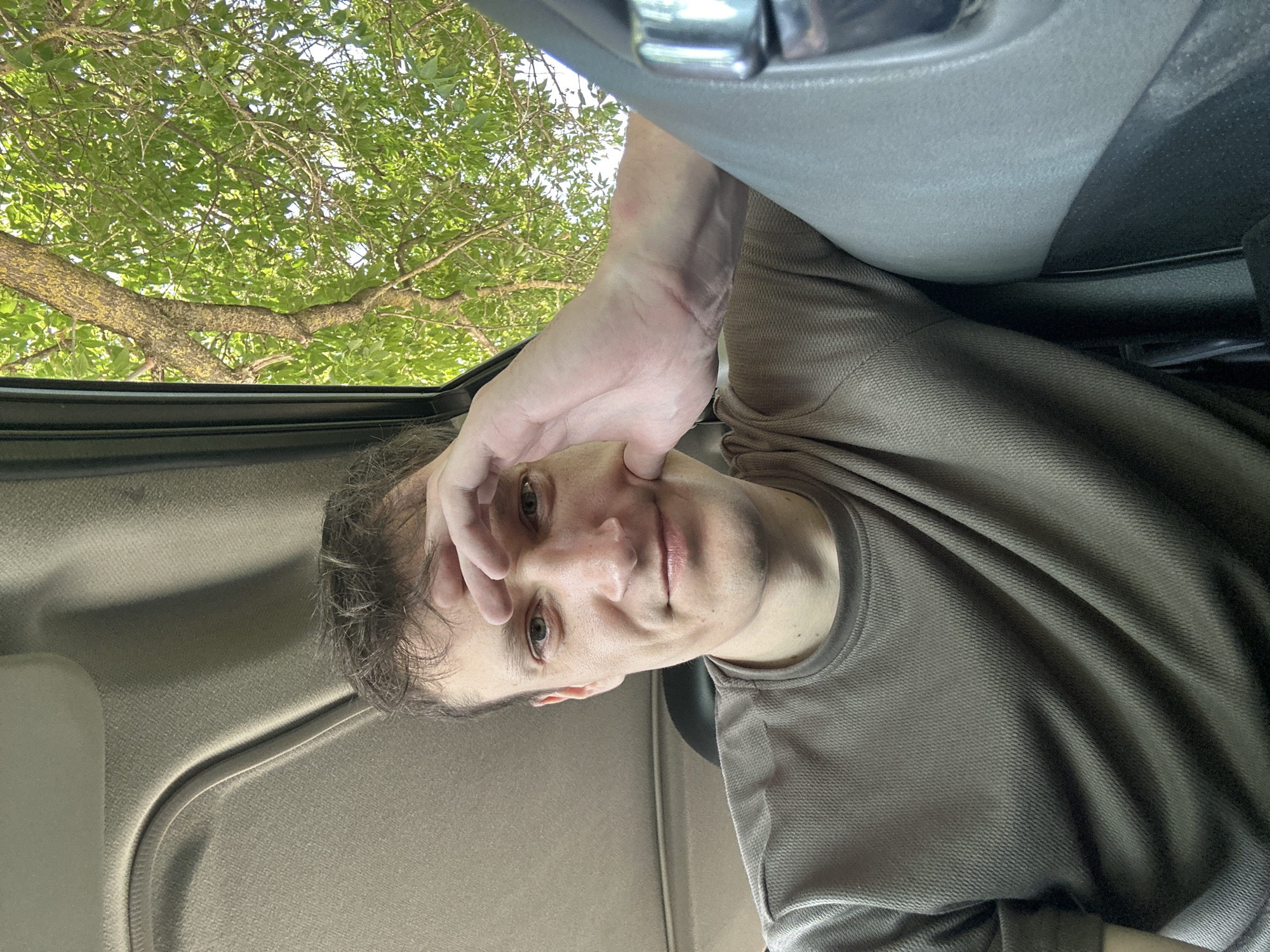 A medium shot is captured from inside a vehicle, looking outwards. A man with light brown, tousled hair and light blue eyes is seated in the car. He has a slight smile and is holding his right hand to his forehead and cheek. He is wearing a brown, textured crew-neck t-shirt. The interior of the car, including the headliner and part of the door panel, is visible. Outside the car window, a tree with green foliage and a textured brown trunk is visible against a bright sky. The scene is set in the city of Makiv, Ukraine.