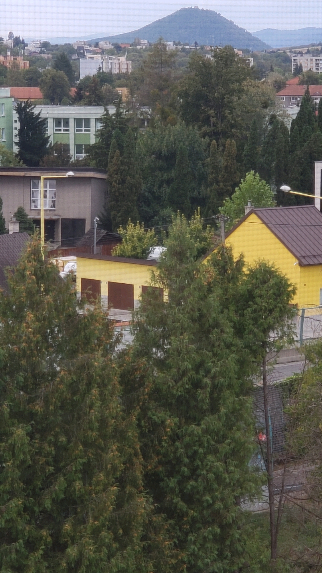 The image captures a view of a residential area in Prešov, Slovakia, with a focus on buildings and vegetation. In the foreground, several coniferous trees are prominent, obscuring parts of the lower section of the scene. Behind them, a bright yellow building with a dark brown roof is visible, featuring two large, dark brown garage doors. Adjacent to this yellow structure, another building with a darker roof and cream-colored walls is partially seen, with a single window and a yellow vertical pole in front of it. Further in the background, a light green building with white framed windows is visible. Beyond these structures, a distant mountain range under a cloudy sky forms the backdrop. The scene appears to be an outdoor, daytime view, likely from an elevated position, looking over rooftops and trees within a suburban or urban environment.