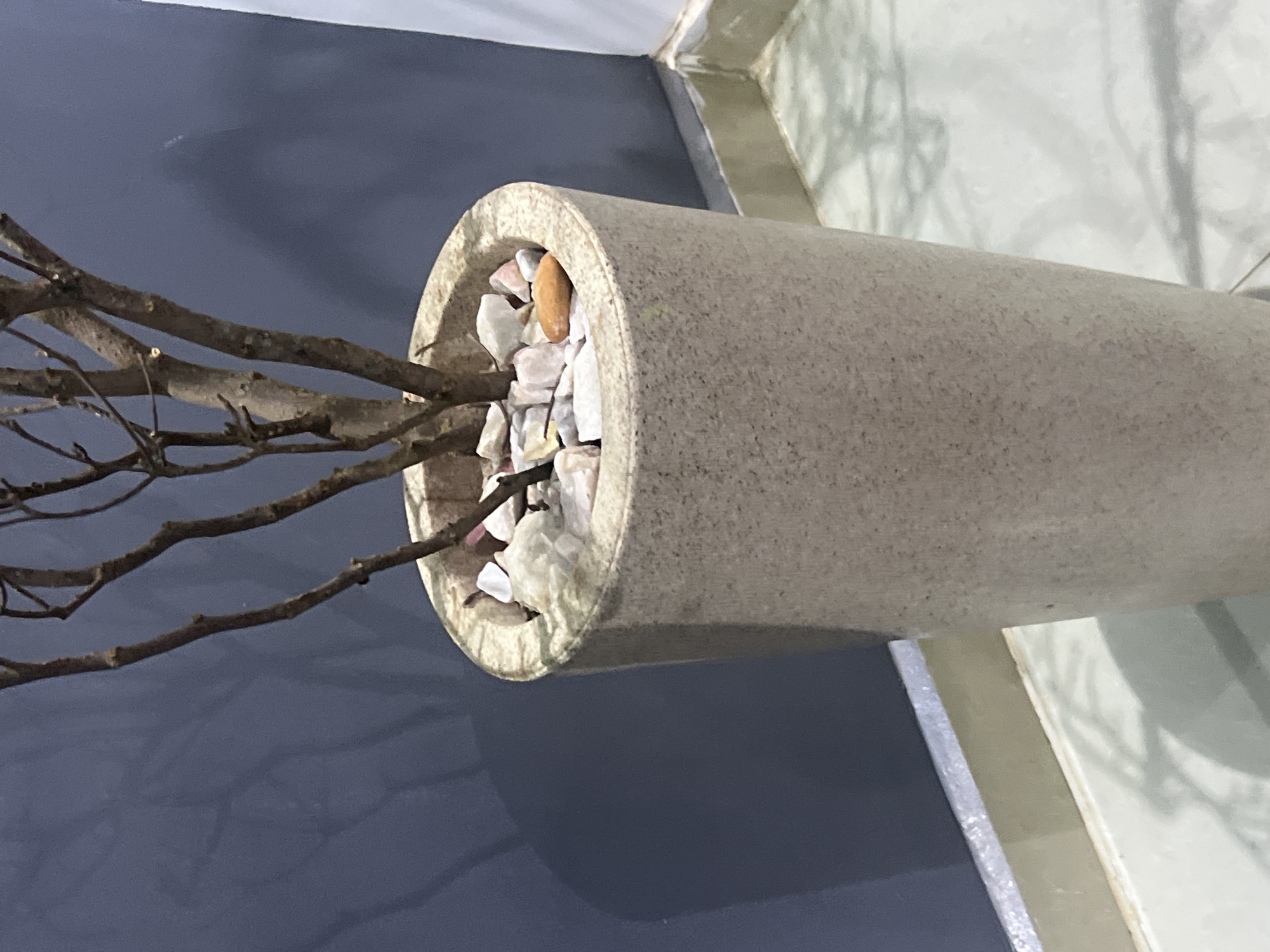 The image shows a tall, cylindrical planter pot made of a speckled, light-colored material. The pot contains a bare-branched plant with thin, brown stems. The surface of the soil within the pot is covered with small, white and pinkish stones, and one larger, oval-shaped orange stone is visible on top. The pot is placed indoors, next to a dark blue wall that casts a soft shadow. The floor appears to be light-colored tiles, with a shadow of the plant's branches cast upon it. A white vertical element is partially visible on the right side of the image. The scene is domestic, likely within a home or office. There are no people or animals visible in the image. The location in Gura Top, Nigeria is not depicted in the image.