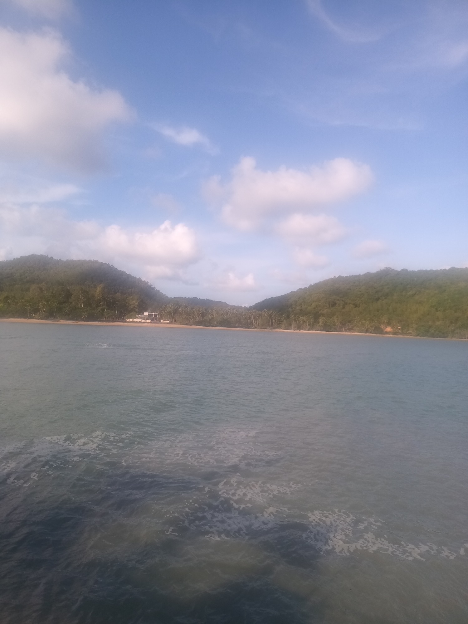 The image depicts a coastal scene in Tambon Hat Sai Ri, Thailand. The foreground is dominated by water, with visible surface movement and some foam. In the middle ground, there is a sandy beach lined with palm trees, behind which rise hills covered with lush green vegetation. A building is discernible on the beach. The sky is light blue with scattered white clouds.