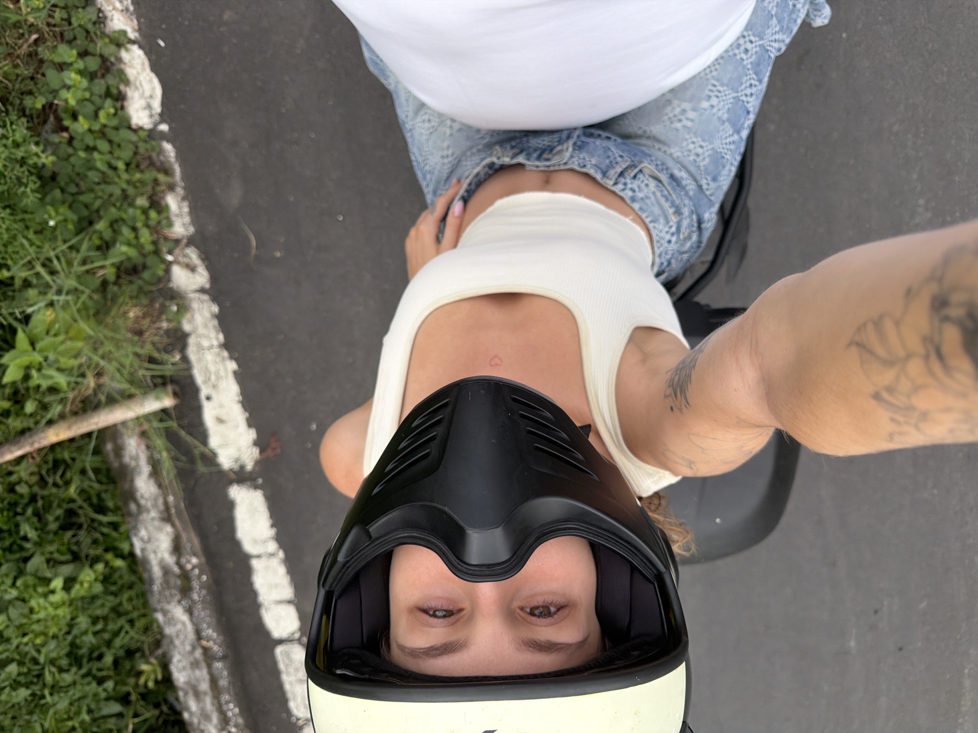 A high-angle, over-the-shoulder shot captures a woman wearing a yellow and black motorcycle helmet with a black face mask, her eyes visible through the opening, looking upwards. She is seated on a motorcycle, wearing a white ribbed tank top and distressed light blue denim jeans. A small red heart tattoo is visible on her chest. A person's tattooed arm is in the foreground, suggesting they are either driving the motorcycle or taking the photo. The background consists of a gray asphalt road bordering a patch of vibrant green foliage and a white concrete curb, indicative of a roadside in North Kuta, Indonesia.