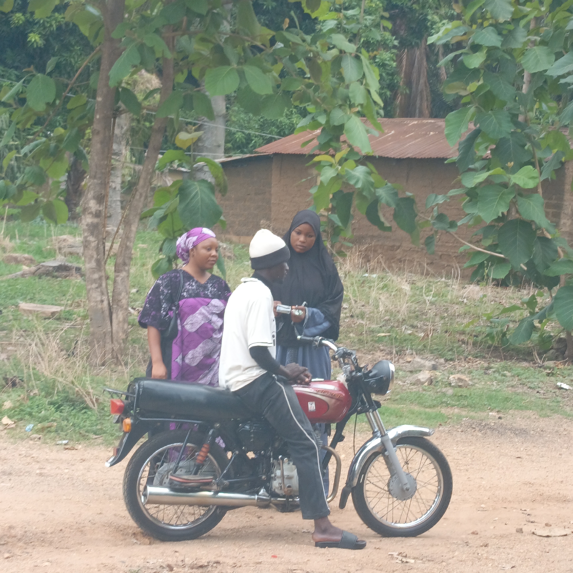 The image depicts three people near a motorcycle. A man is sitting on the motorcycle wearing a white shirt, dark trousers with a white stripe, a black and white hat, and sandals. To his left stands a woman in a purple patterned dress and headscarf. To the right of the man, another woman stands wearing a black hijab and a blue dress. They appear to be conversing. The motorcycle is red with chrome accents. The background shows a dirt road, trees, and a mud brick building with a corrugated metal roof. The location appears to be a rural or semi-rural setting in Andaha, Nigeria.