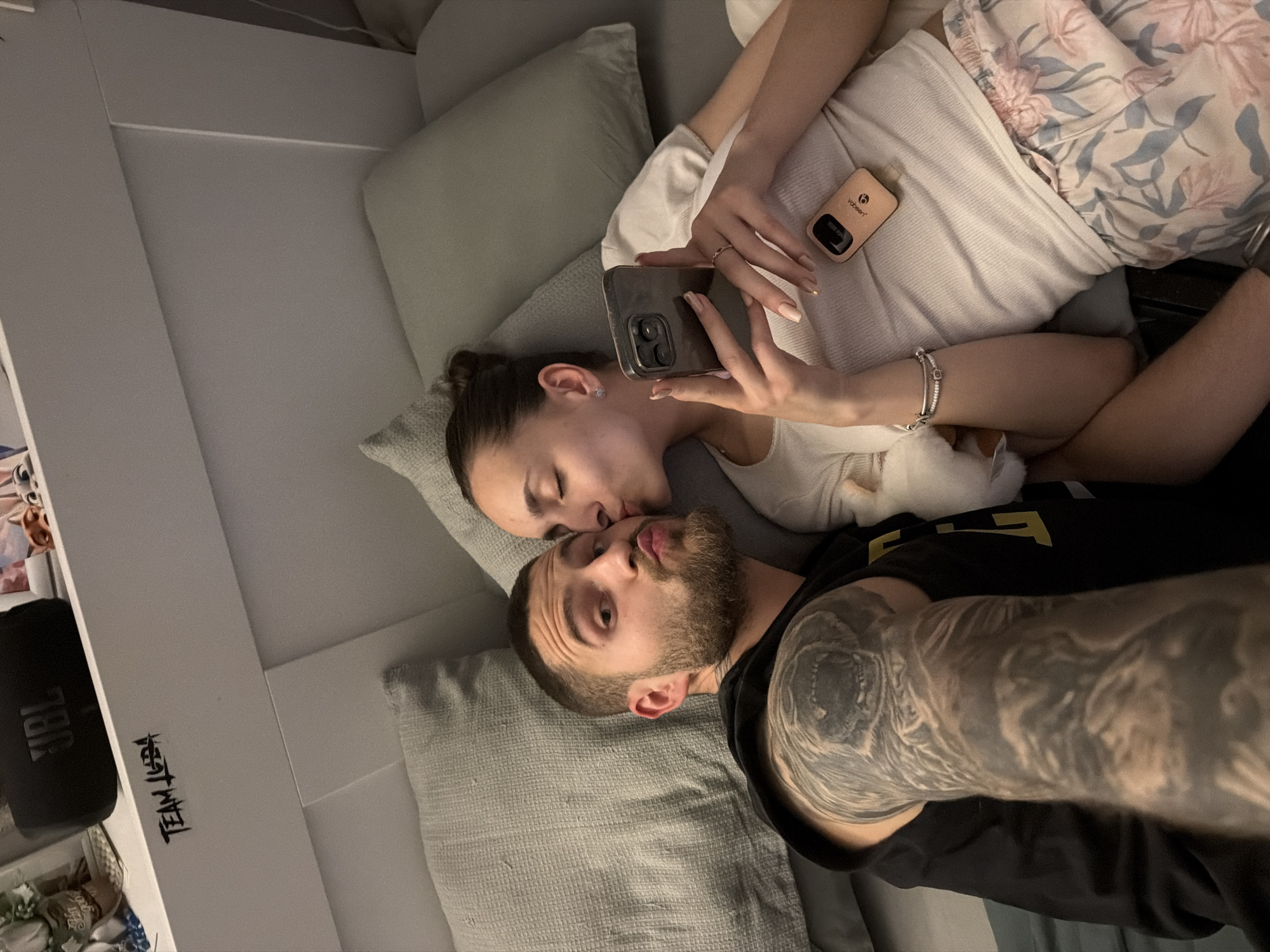 This intimate overhead shot, taken in Sofia, Bulgaria, captures a couple relaxing indoors, likely on a bed or couch. The man, with a beard and a heavily tattooed arm (a full sleeve featuring a detailed design, possibly a skull or creature's head on the bicep), is lying on his back, looking towards the camera with a playful pout. He wears a black t-shirt. The woman, with her hair tied back, lies beside him, gently kissing his cheek. She is holding a dark-colored smartphone, presumably taking the picture, and wears a white top with floral-patterned shorts. Her hand shows manicured nails and a ring, and she wears a silver bracelet on her wrist.
The setting is casual and affectionate, with grey pillows and bedding. In the background, to the left, a black JBL speaker is visible on a surface, next to a white panel that has "TEAM LEBEN" written vertically in black, stylized script. Another small beige electronic device, possibly a vape or charger, with the brand "Vabeen" visible on it, rests next to the woman's side. A small white pet, possibly a cat or dog, is partially visible near the man's arm and the woman's waist. The lighting suggests it is evening or nighttime.