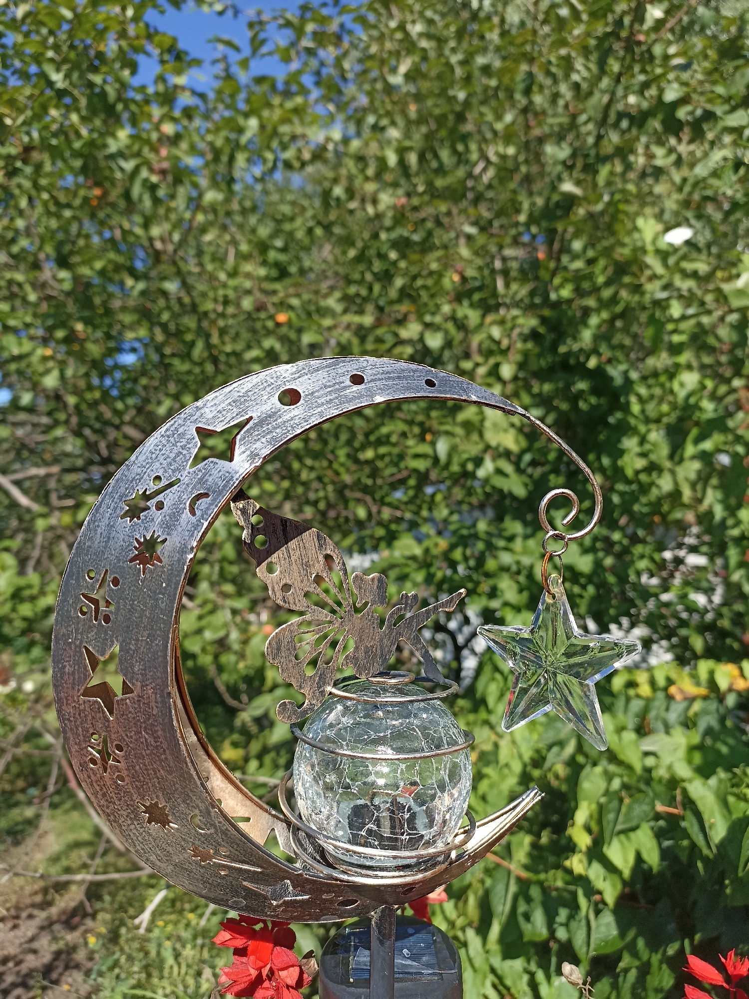 A metallic crescent moon garden ornament is prominently displayed outdoors, set against a backdrop of lush green foliage and a clear blue sky. The ornament is intricately designed with cutouts of stars, moons, and a small fairy. A clear glass sphere, likely a solar light, rests within the curve of the moon. Hanging from a delicate wire loop attached to the top of the moon is a faceted glass star, catching the sunlight. The ornament is mounted on a dark-colored pole, partially visible at the bottom of the frame. Red flowers are visible at the base of the pole. The scene appears to be in a garden setting, possibly in the city of Mochische, Russia, given the lush vegetation and daytime lighting. No people or animals are present in the frame.