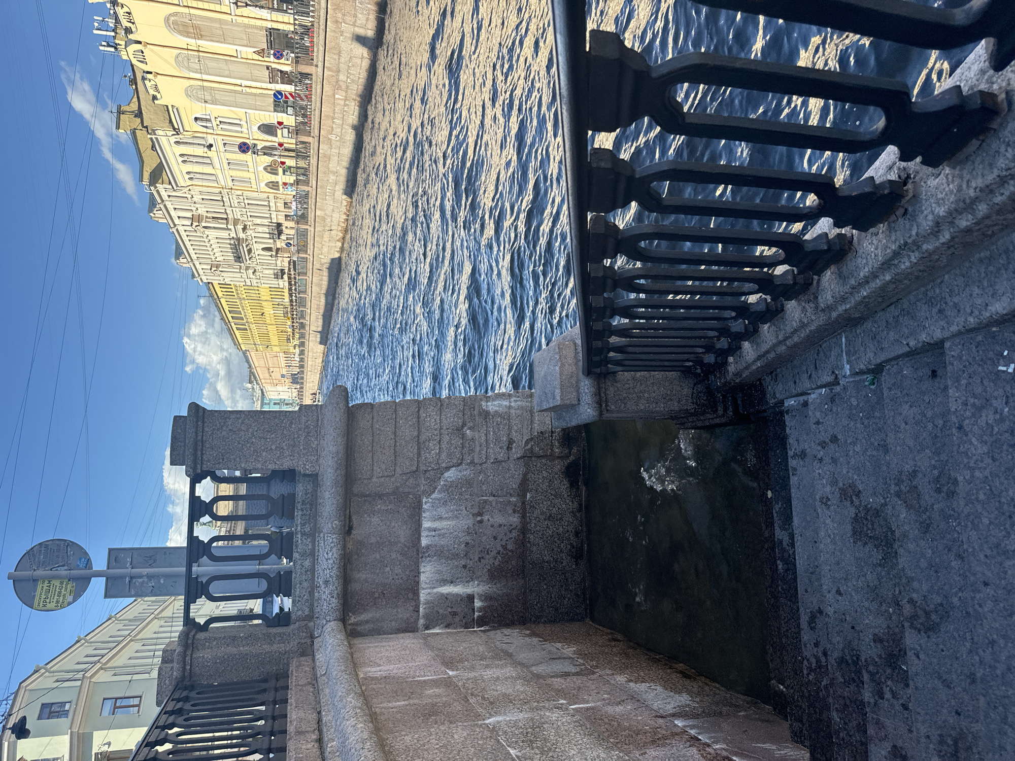 The image captures a scene along a canal in Saint Petersburg, Russia, likely the Griboyedov Canal given the architectural style and waterway. In the foreground, a granite embankment features a dark metal railing with decorative, oval-shaped openings. Stone steps descend from the embankment to the water's edge, where a small pool of dark water is visible, disturbed by ripples. The embankment wall is constructed from large granite blocks.
Beyond the embankment, the canal stretches into the distance, its blue water reflecting the bright sunlight. Along the opposite bank, a row of buildings displays a uniform architectural style, characterized by pastel yellow facades, numerous windows, and ornate detailing, suggesting a historic urban setting. A blue sky with scattered white clouds forms the backdrop, and power lines are visible overhead.
A circular sign on a pole stands near the top of the embankment, featuring text in Cyrillic, indicating it is likely informational or directional. The scene is devoid of people. The lighting suggests it is daytime, with the sun casting shadows and highlighting the textures of the stone and water. The overall impression is one of a serene, sunlit urban waterway.