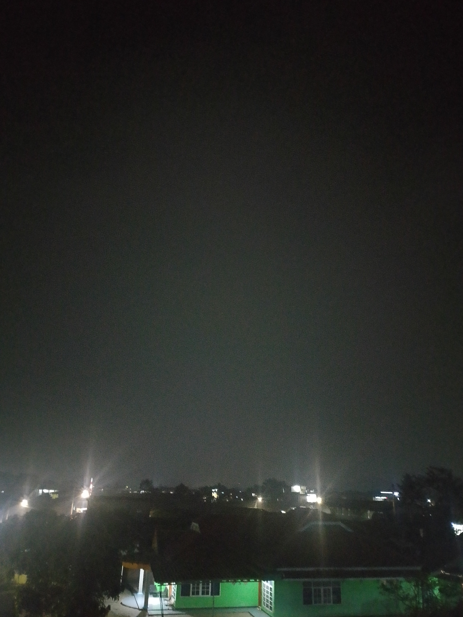 A nighttime aerial view captures a dark, hazy sky over a residential area. In the foreground, a bright green building with white-framed windows is visible, along with a covered porch area and some trees. Beyond this, a collection of rooftops and other buildings are dimly illuminated by distant lights. Further in the distance, more structures and lights are scattered across the landscape, suggesting a populated urban or semi-urban environment. The overall impression is one of a quiet, dimly lit night in Situwangi, Indonesia.