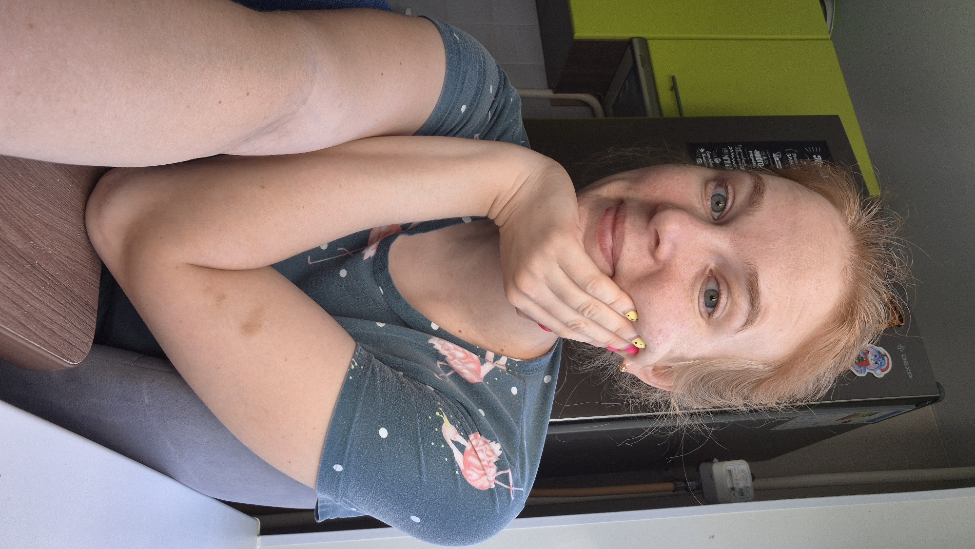 A woman with red hair and blue eyes is posing for a selfie indoors. She is wearing a blue shirt with white polka dots and pink flamingoes. Her right hand is covering part of her face. Her fingernails are painted pink with yellow chick designs. She is sitting at a brown table with a white surface underneath. Behind her is a refrigerator with a Smurf sticker on it, and a green kitchen cabinet. The location is Chaikovsky, Russia.