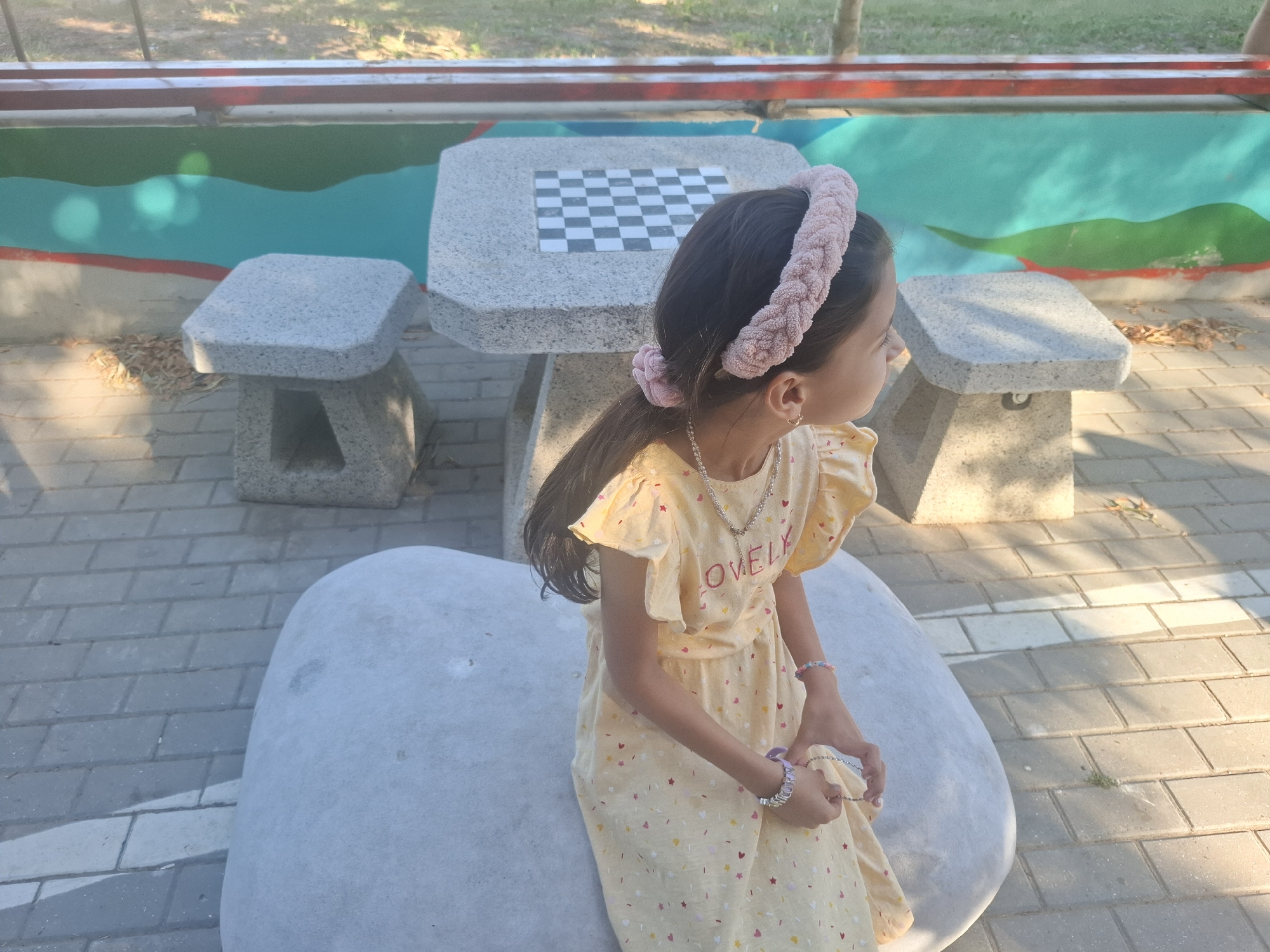 In an outdoor setting in Vadul lui Voda, Moldova, a young girl is seated on a large, light gray, pebble-shaped bench. She has long, dark hair pulled back with a textured, light pink headband and a matching hair tie. She is wearing a light yellow dress adorned with small red and yellow heart-shaped confetti. A silver necklace with small heart charms rests on her neck, and she is wearing several bracelets on her wrists, including one with purple beads. The girl is looking to her right.
Behind her, there is a gray stone table with a built-in checkerboard. Two matching stone stools are placed to the sides of the table. The ground is paved with light gray rectangular bricks. In the background, a low wall features a painted mural with wavy bands of green, turquoise, and red. Beyond the wall, a patch of grass and trees is visible under a sunny sky. The lighting suggests it is daytime.