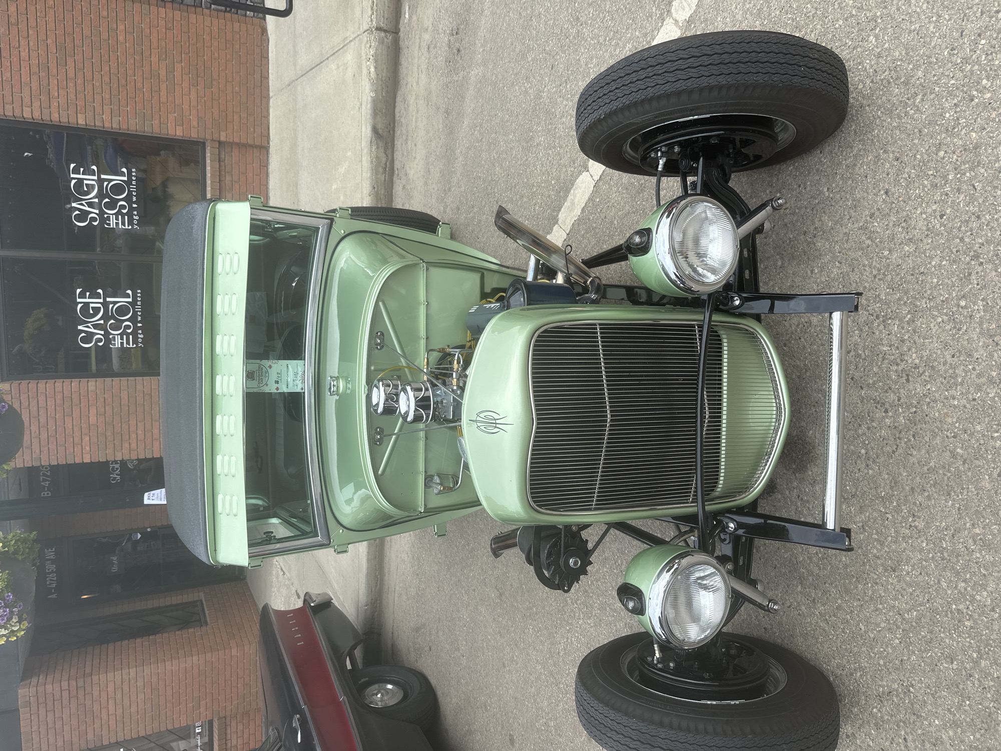 A light green 1930s-style hot rod is parked on an asphalt surface, possibly a street or parking lot, in front of a brick building. The building has large windows with "SAGE the SOL yoga & wellness" signage. A black classic car is partially visible to the left of the hot rod. The hot rod features an exposed engine with chrome detailing, a vertical grille, and prominent headlights. A small award or plaque with the text "BEST IN CATEGORY Vintage" is affixed to the windshield. The scene appears to be outdoors, with a concrete curb and sidewalk visible. The setting is the City of Leduc, Canada, indicated by address numbers "A-4726 50th AVE" and "B-4726" on the building.