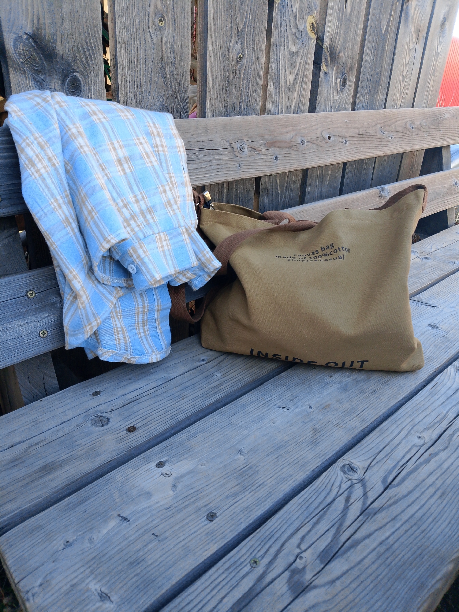 The image shows a light blue and tan plaid shirt draped on the left side of a weathered wooden bench. To the right of the shirt is a tan canvas bag with brown straps. The bag has text printed on it that reads "canvas bag made of 100% cotton simple casual" and "INSIDE OUT." The bench is made of gray-blue planks with visible wood grain and screw heads. In the background, there is a fence made of vertical wooden planks, and a glimpse of something red is visible behind the fence on the right. The location is not possible to identify based on the image alone.