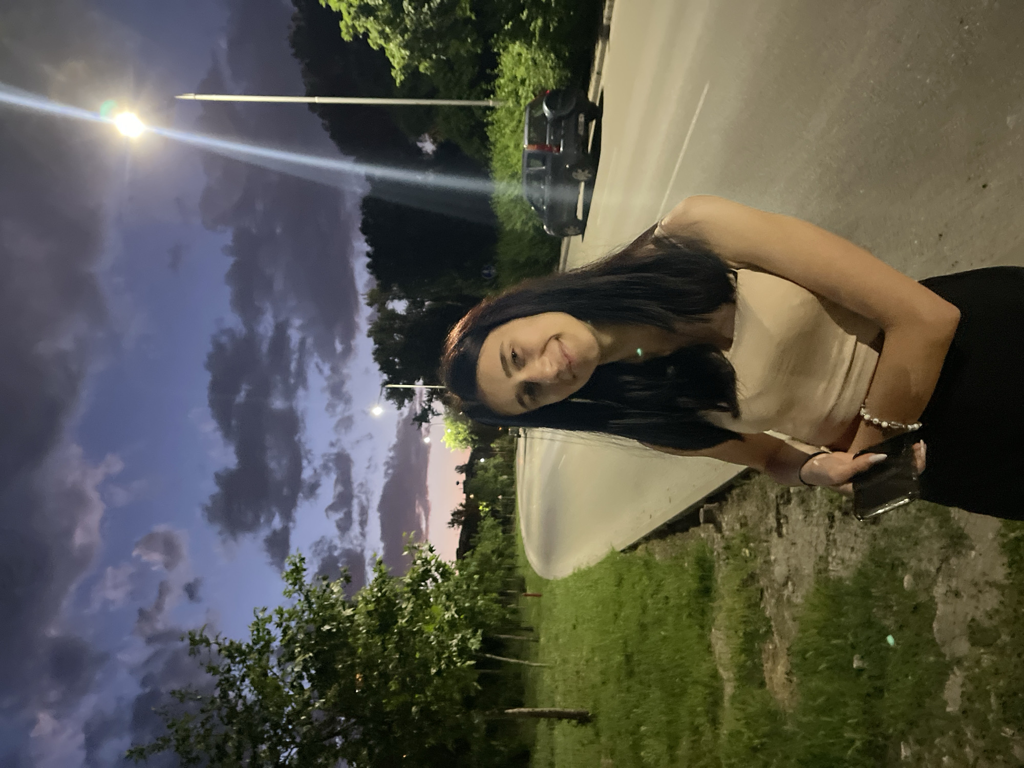 A woman with long dark hair, wearing a light-colored top and dark pants, stands on the side of a road in an urban environment. She is holding a cell phone. Behind her is a grassy area with trees and a car parked on the roadside. The sky is filled with dark clouds and there is a street lamp illuminating the scene.