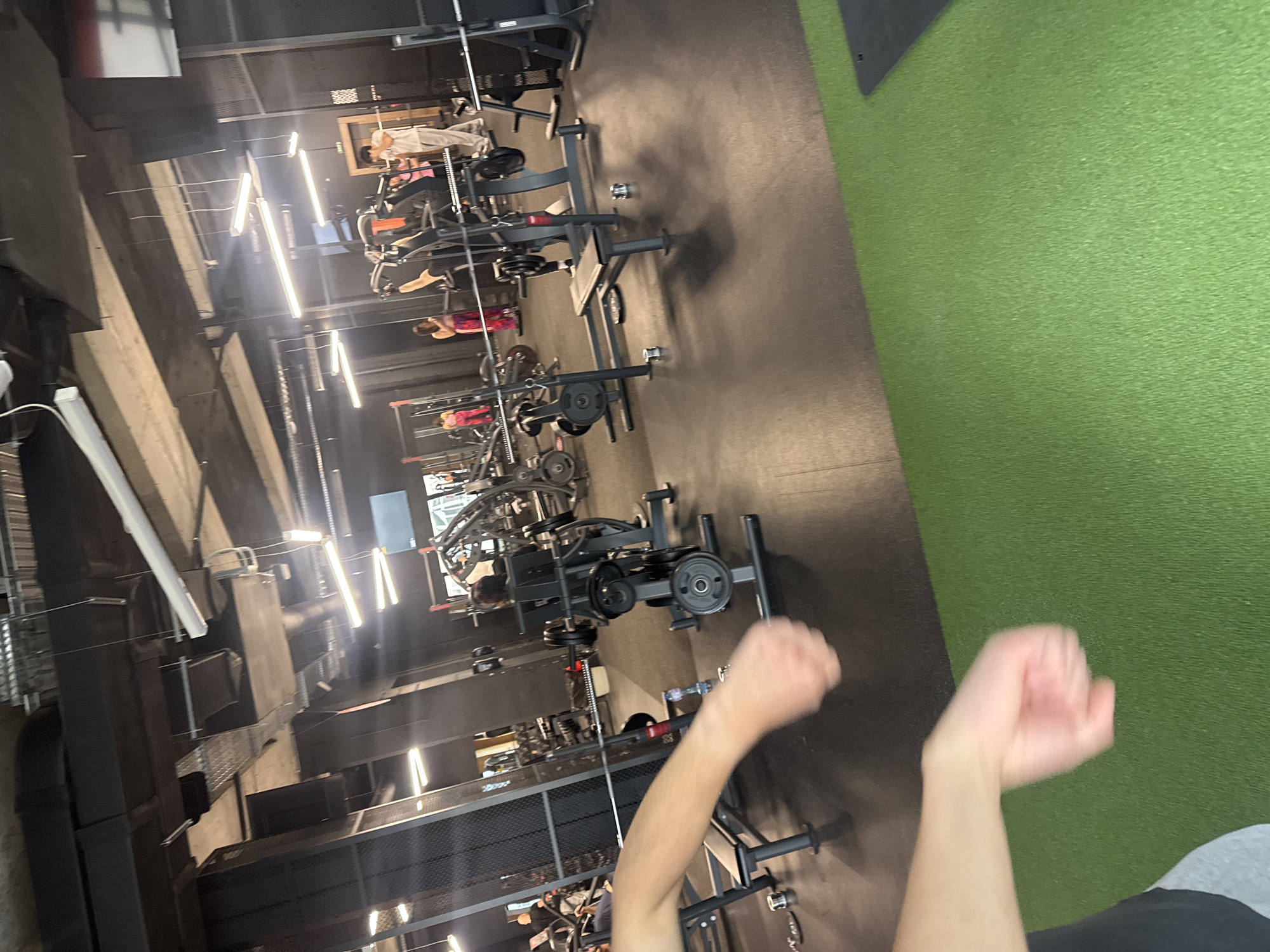 This vertically oriented image captures an interior view of a modern gym or fitness facility, taken from a low perspective, likely from someone lying or seated on the floor. In the immediate foreground, a pair of light-skinned arms with partially clenched fists are visible, indicating the photographer's position.
The gym floor features a dark, possibly rubberized surface that transitions into a vibrant green artificial turf area on the right side of the frame. Numerous pieces of black and grey weightlifting equipment, including benches, plate-loaded machines, and barbells, are arranged in rows across the darker flooring. Several individuals are present, some actively using the equipment. One person wearing a white top is visible in the upper right, seated on a machine. Further down, another individual in a pink or red top is also engaged with equipment.
The ceiling reveals an industrial aesthetic with exposed concrete beams, metal grating, and prominent ventilation ducts. Multiple bright, linear fluorescent light fixtures illuminate the space, creating reflections on the darker surfaces. The overall setting suggests a functional and well-equipped workout environment. There is no discernible text or branding visible on any objects or equipment. The time of day cannot be determined from the interior lighting.