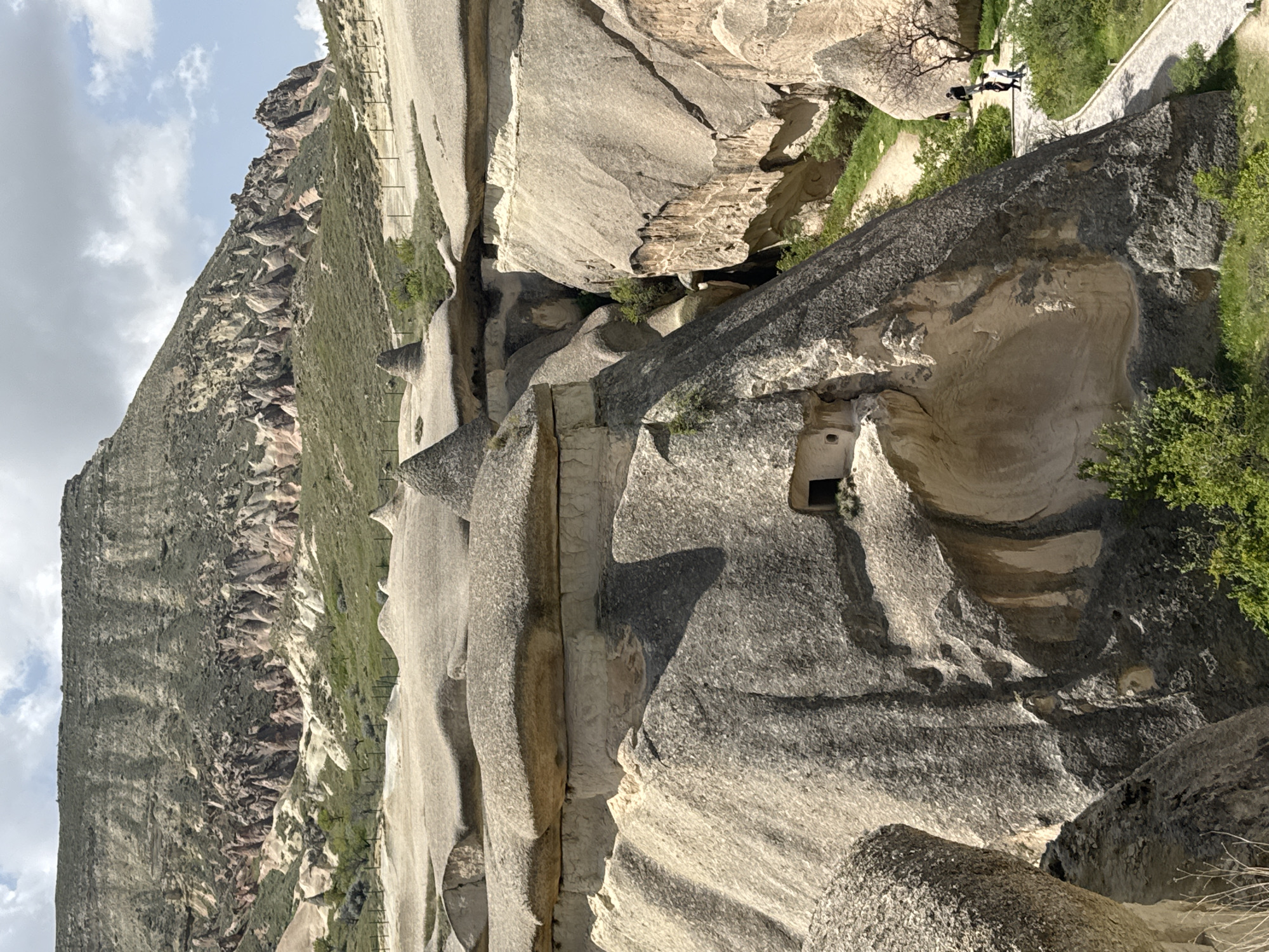 This image depicts a portion of Cappadocia's unique landscape, likely within the city of Avanos, Turkey. The scene is dominated by eroded rock formations, characterized by their soft hues of tan, gray, and cream. The geological features include caves, carved openings, and naturally sculpted shapes. A section of green landscape featuring an eroding rocky wall is partially visible to the left. A pathway is seen at the upper right, with two individuals visible upon it. The day appears overcast with patches of blue sky and white clouds.