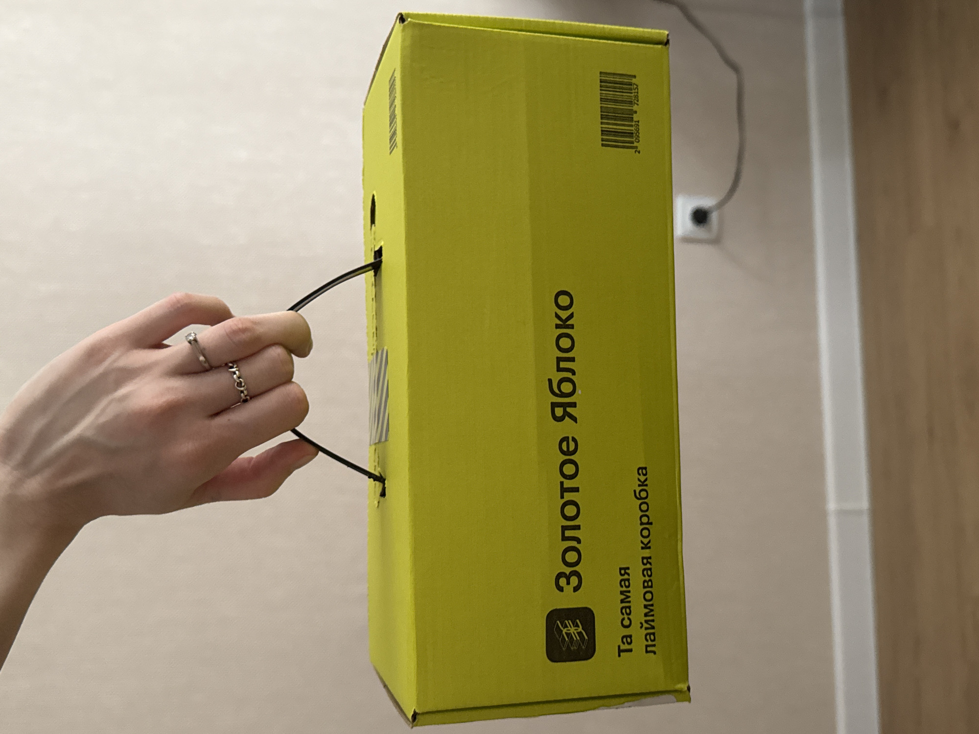 A hand with a ring is holding the handle of a bright yellow box. The box has text in Russian, which translates to "Golden Apple" and "The most lime box." There is also a barcode on the side of the box. The background is a plain wall. This image does not contain any information about the city of Perm, Russia.