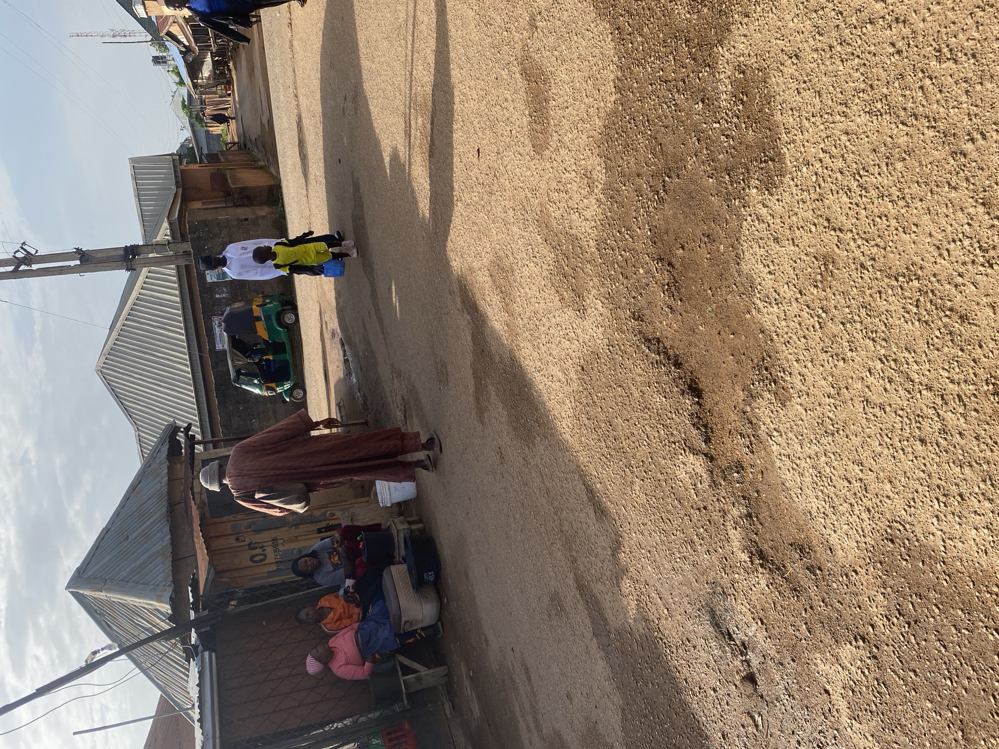The image depicts a street scene in the city of Jos, Nigeria, characterized by a dry, dusty, and uneven road surface with visible potholes. On the left side of the road, a shaded area under a corrugated metal roof houses several people. Three women are seated on a bench, with one appearing to be the central figure, wearing an orange top and dark pants, engaged in conversation with a woman in a pink headscarf and a blue garment. Another woman in a grey t-shirt is also present. Beside them, a man in a brown robe and a light-colored cap walks away from the camera, carrying a bag over his shoulder and holding a cane. In the background, a green and yellow auto-rickshaw is parked. Further down the street, a man in a white shirt and a child in a yellow shirt and shorts are walking away from the viewer. The architecture suggests a blend of informal and semi-permanent structures with corrugated metal roofing. The sky is partly cloudy, indicating a daytime setting. The overall impression is that of a typical, bustling street in a West African city, with everyday life unfolding in a somewhat arid environment.
