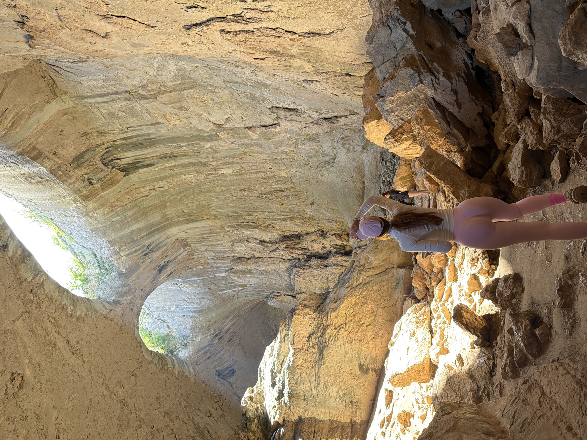 The image shows a woman standing in Devetashka Cave near Karlukovo, Bulgaria. She is wearing pink leggings, a light-colored long-sleeved shirt, a pink hat, and is facing away from the camera. The cave has large openings to the outside, visible in the image. The interior is characterized by layered rock formations and rough, rocky surfaces.