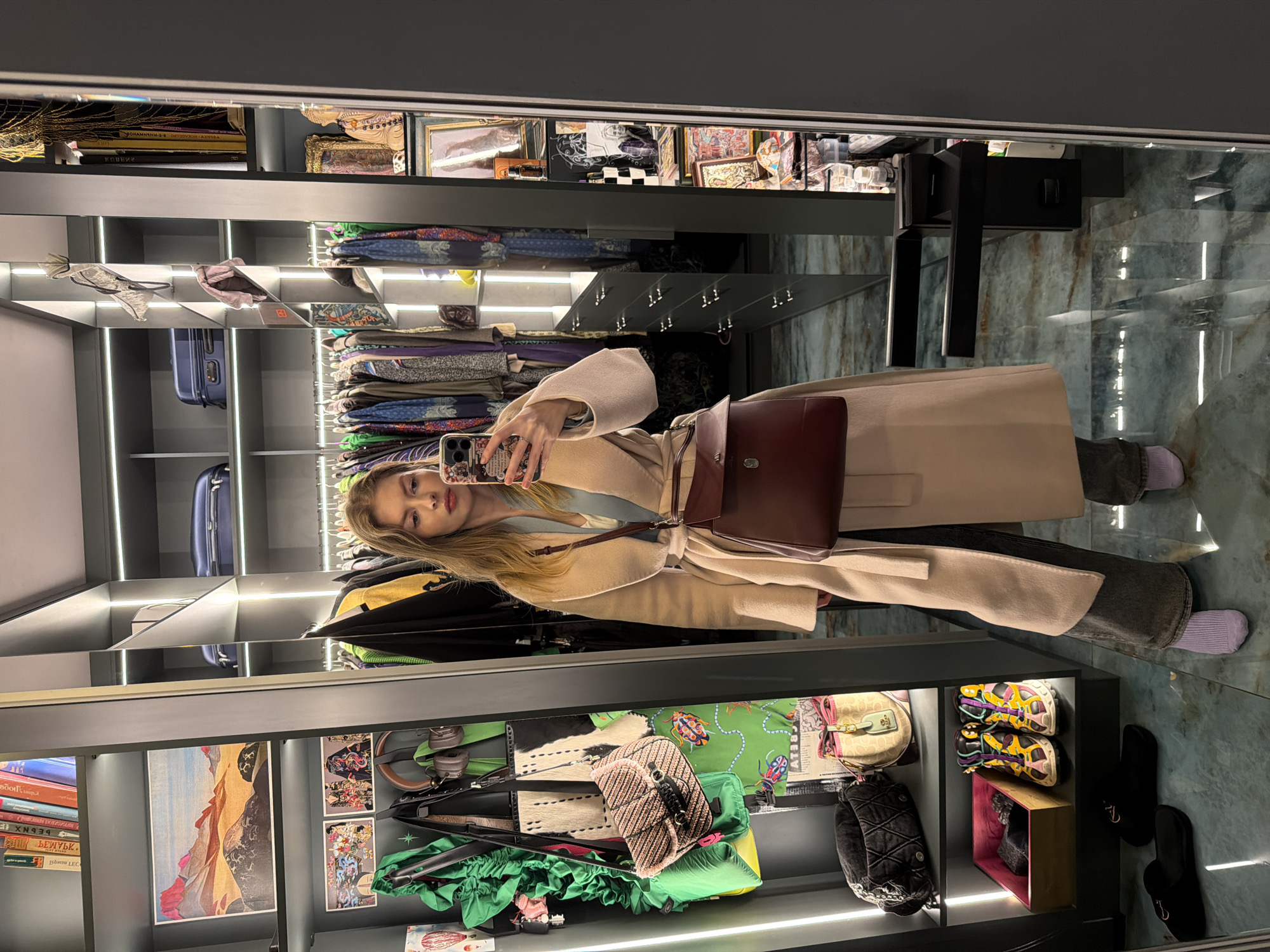 A person is taking a selfie in what appears to be a walk-in closet. The closet has shelves filled with various items including books, artwork, bags, clothes, and shoes. There are hanging clothes visible, and some luggage on a shelf. The floor has a marbled pattern. The person is wearing a light-colored coat and carrying a brown bag. The environment is well-lit. There is no other information to suggest this image has any connection to the city of Moscow, Russia.