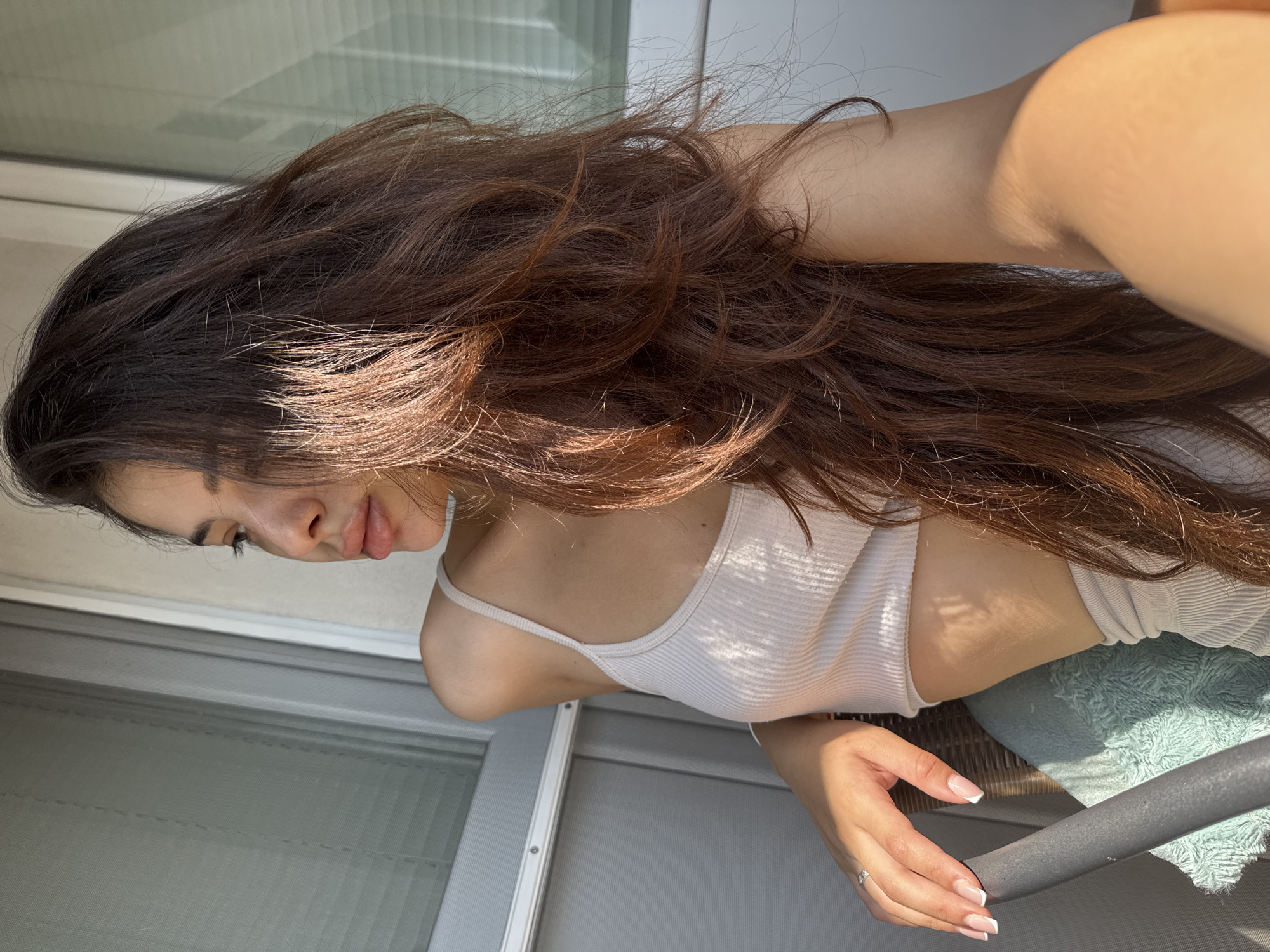The image depicts a young adult female positioned on an outdoor balcony in Plovdiv, Bulgaria. She is seated, facing her left in profile, with her gaze directed horizontally towards the left side of the frame. Her long, dark brown hair, with visible lighter auburn tones where struck by direct sunlight, flows over her shoulders and back. She wears a light beige ribbed crop tank top and matching ribbed high-waisted bottoms. A silver ring is present on her left ring finger, and her fingernails display a French tip manicure. Her left forearm rests on the dark grey padded armrest of a woven chair, which features a light teal, textured cushion. Strong, direct sunlight from the right casts prominent shadows across her body, hair, and the chair cushion. The background consists of a white-framed window or sliding glass door with horizontal blinds to her left, and another similar white-framed door to her right.