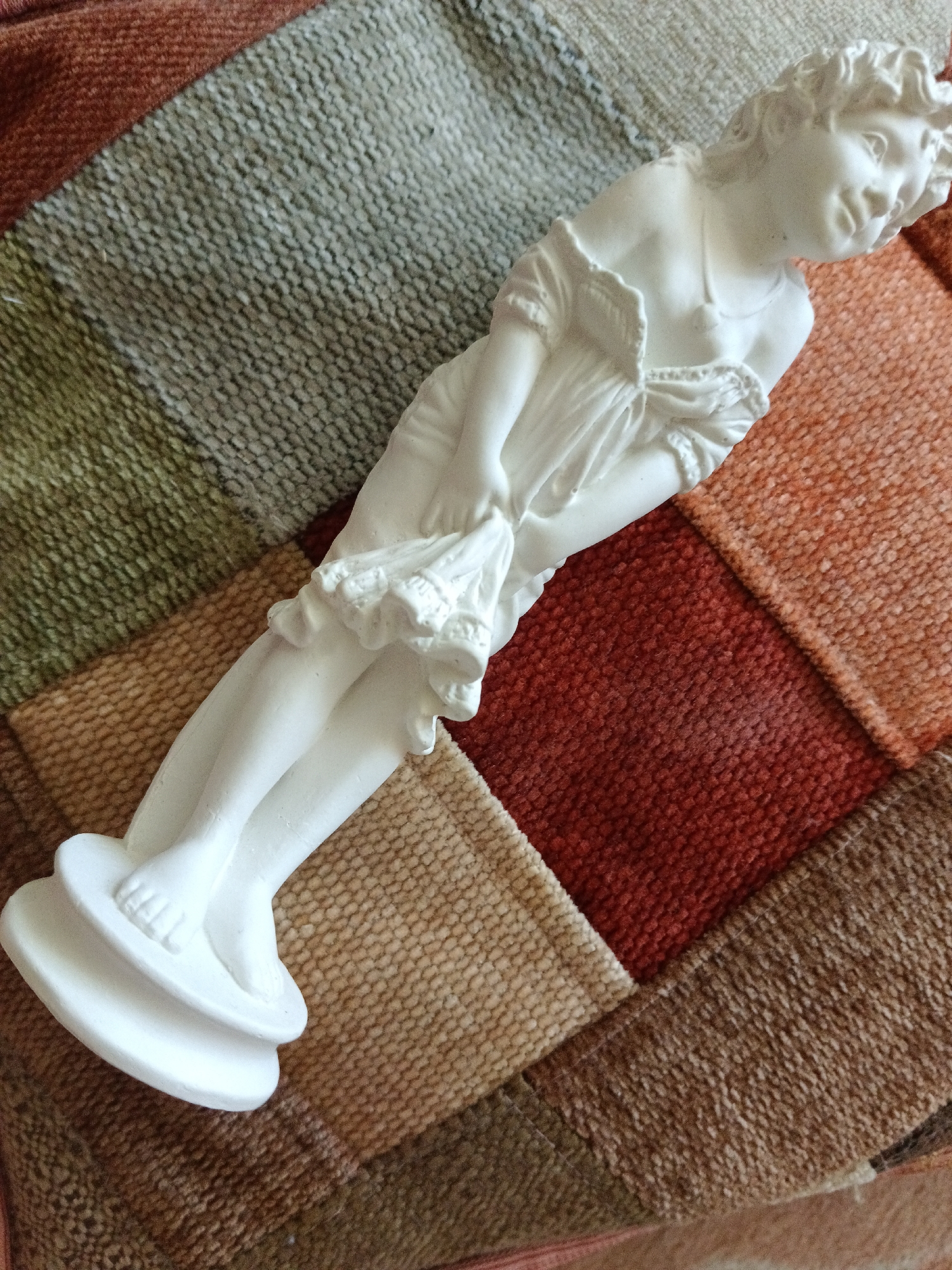 A white statuette of a young woman in a flowing dress stands on a multi-colored patchwork textile. The textile has squares of varying shades of brown, green, and red. The statuette is positioned diagonally across the textile, with its feet at the bottom left and its head angled towards the upper right. The woman is depicted in a graceful pose, holding a piece of fabric in her hands. The statuette is smooth and detailed, with delicate features and clothing. The lighting in the image is natural, casting soft shadows and highlighting the texture of both the statuette and the textile. The scene is indoors, likely in a home setting, but the specific location in Severnyi, Russia, cannot be determined from the image itself.