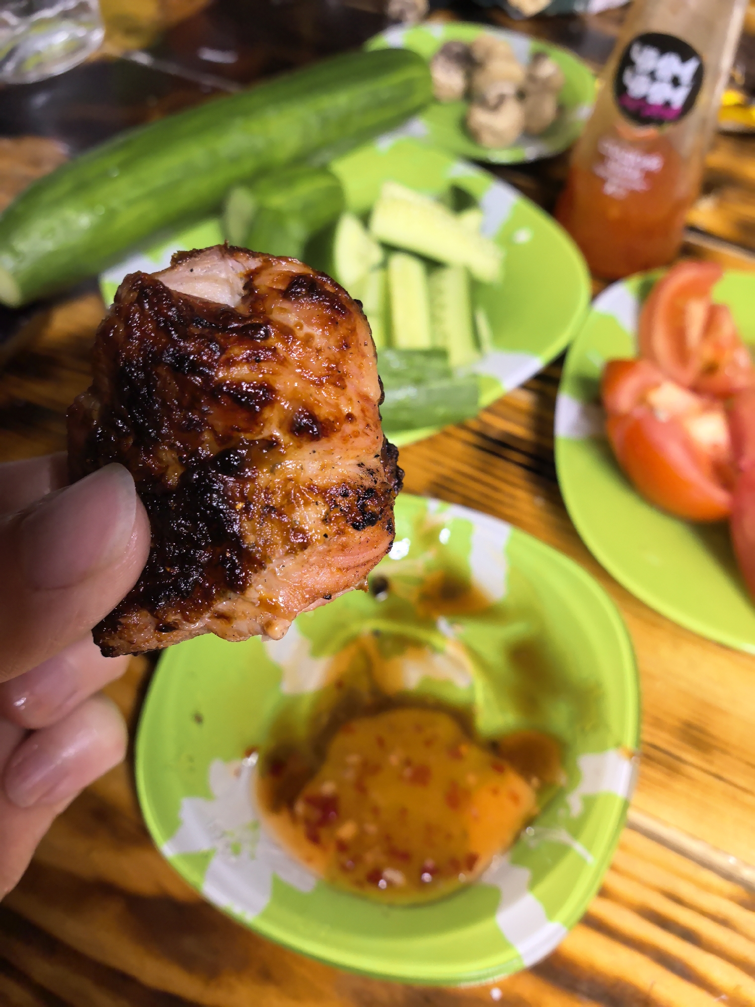 The image captures a close-up, overhead perspective of a person's left hand holding a piece of grilled salmon over a wooden table laden with various food items. The salmon, held between the thumb and forefinger, displays a seared, dark brown and reddish-orange exterior with distinct charring and grill marks. A portion of the cooked fish reveals a flaky, pinkish-orange interior where a bite has been taken. The fingernails of the hand are neatly trimmed and light in color.
Directly below the hand, slightly out of focus, rests a small, bright green bowl featuring a white floral pattern. This bowl contains a viscous, orange-red dipping sauce visibly flecked with chili flakes or spices.
On the wooden table, characterized by its warm brown tones and visible grain, several other dishes are presented on identical bright green plates with white floral motifs. To the upper left, a long, dark green whole cucumber is partially visible. Further back, another green plate holds a quantity of uniformly cut green cucumber sticks. To the right, a third green plate displays multiple wedges of red tomatoes. In the background, a small, indistinct green plate contains several small, round, light brown items, possibly mushrooms or small potatoes. Behind the tomato plate, a translucent amber-colored bottle with a dark label, featuring white and pink graphics, is visible, containing an orange liquid. The scene is set indoors in the city of Гаревая, Russia.
