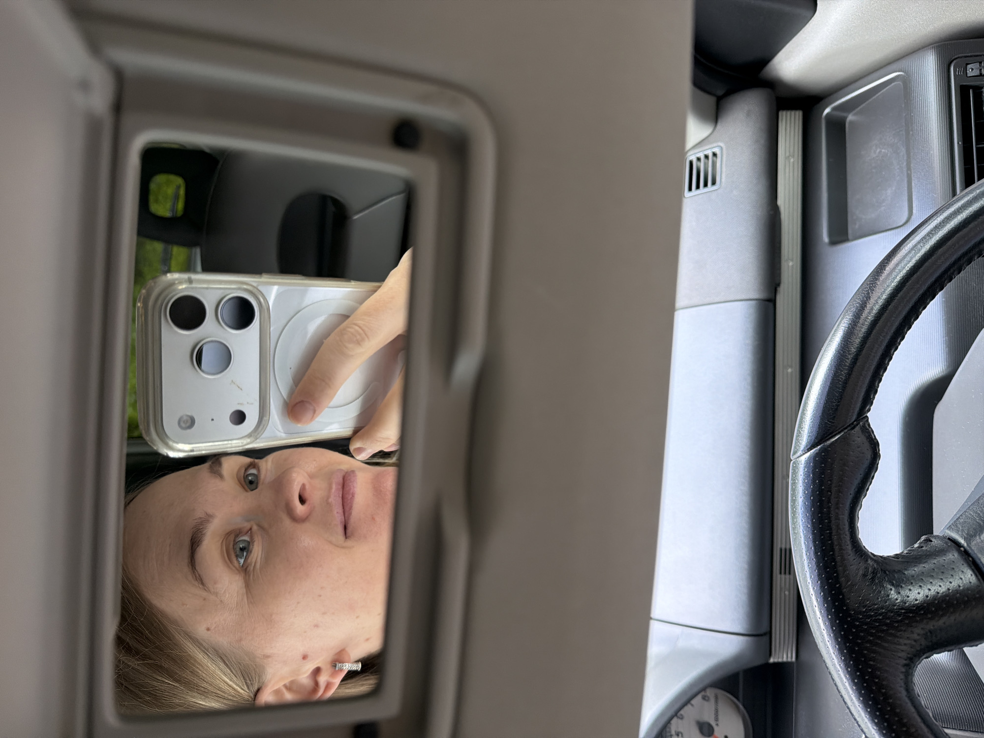 A woman is taking a selfie in a car's vanity mirror. She has blonde hair and is wearing a silver earring. She is holding a smartphone in her right hand, which is also reflected in the mirror. The background shows the interior of a car, including a steering wheel and dashboard. There is no indication of the city of Khabarovsk, Russia, or any specific location.
