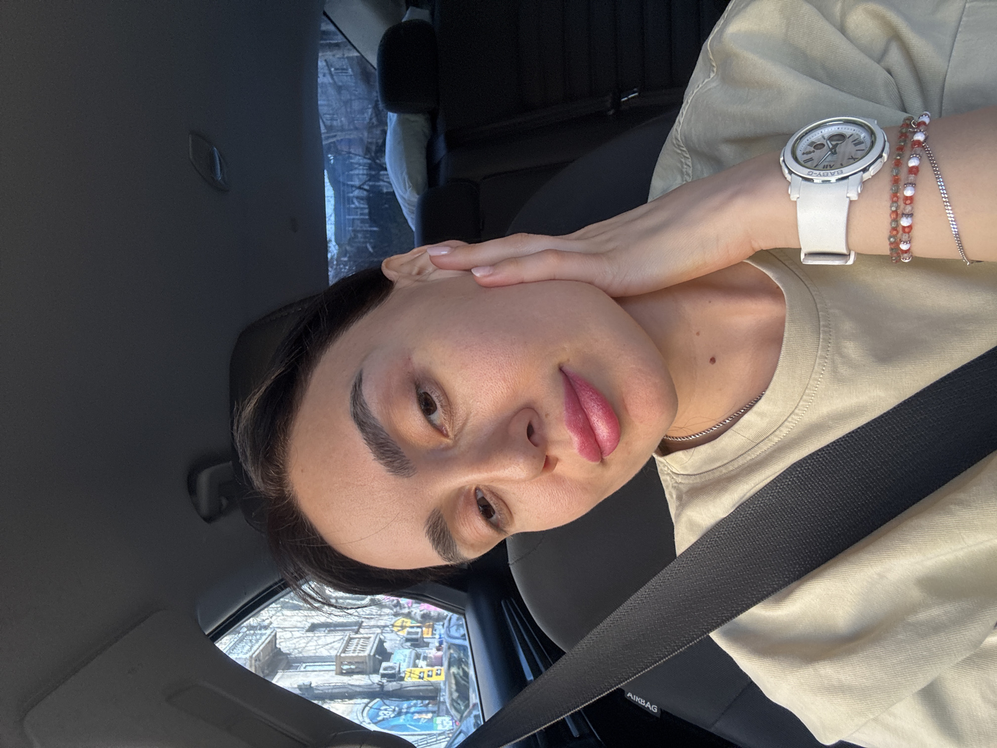 A young adult woman sits in the driver's seat of a car, wearing a seatbelt and a tan t-shirt. She has dark hair, prominent eyebrows, and pink lipstick. She is wearing a white Baby-G watch and bracelets on her left wrist, and her left hand is touching her face near her ear. The background visible through the car windows shows a street scene with buildings, cars, and graffiti, indicative of an urban environment. The location is Tbilisi, Georgia, based on architectural styles, graffiti, and the general atmosphere of the urban landscape. The woman is looking towards the camera with a slight smile.