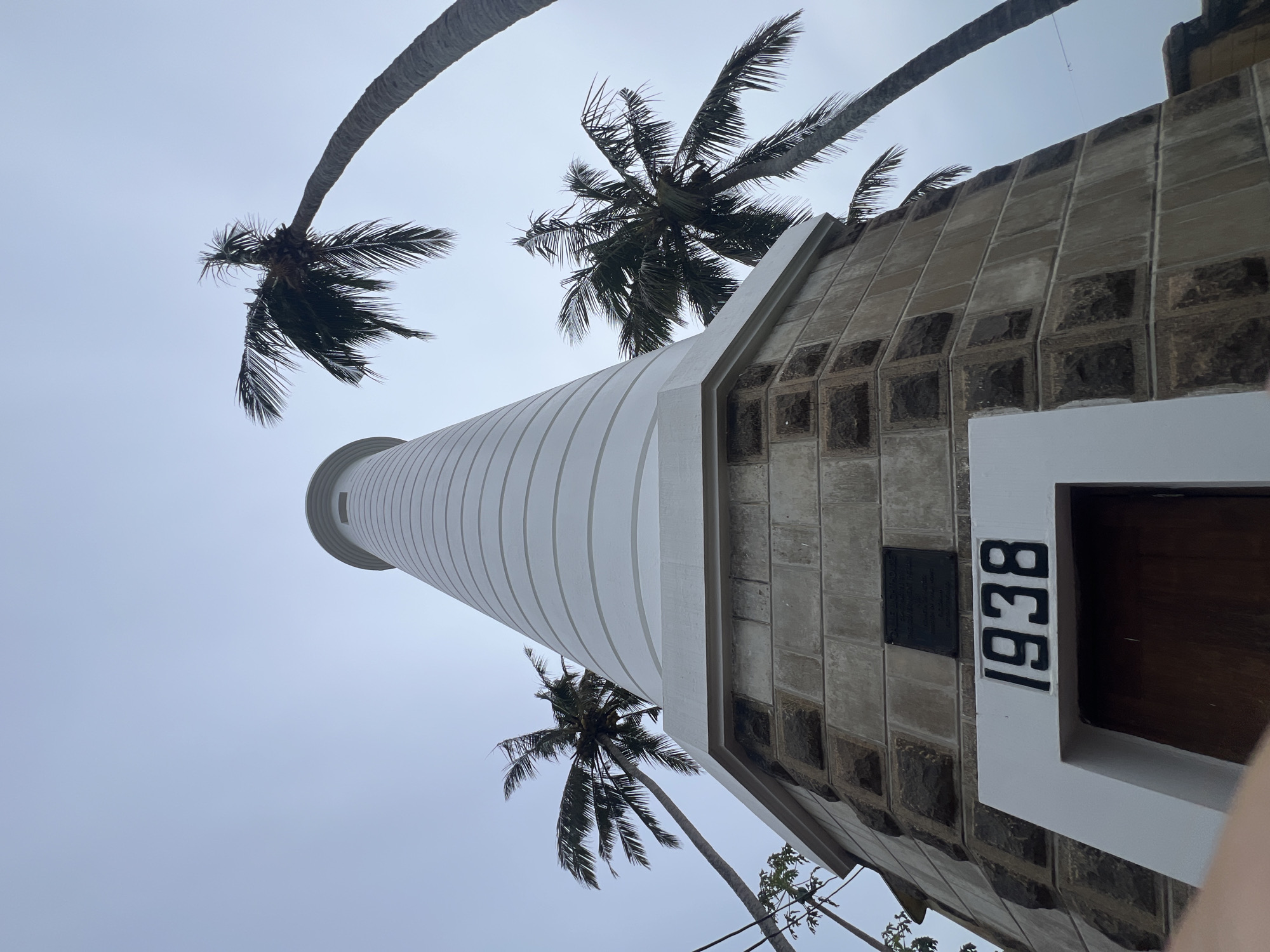 The image shows the Galle Lighthouse in Galle, Sri Lanka, viewed from a low angle looking up. The lighthouse is painted white and has horizontal banding. Its base is made of stone blocks. The year "1938" is prominently displayed in black numbers above a dark wooden door. A plaque is visible on the stone portion of the lighthouse, but its text is illegible. Several palm trees with lush fronds surround the lighthouse, with their trunks leaning at various angles. The sky is overcast and appears a pale blue-grey. Electrical wires are visible in the upper left corner of the image. The scene suggests a slightly humid, tropical environment.
