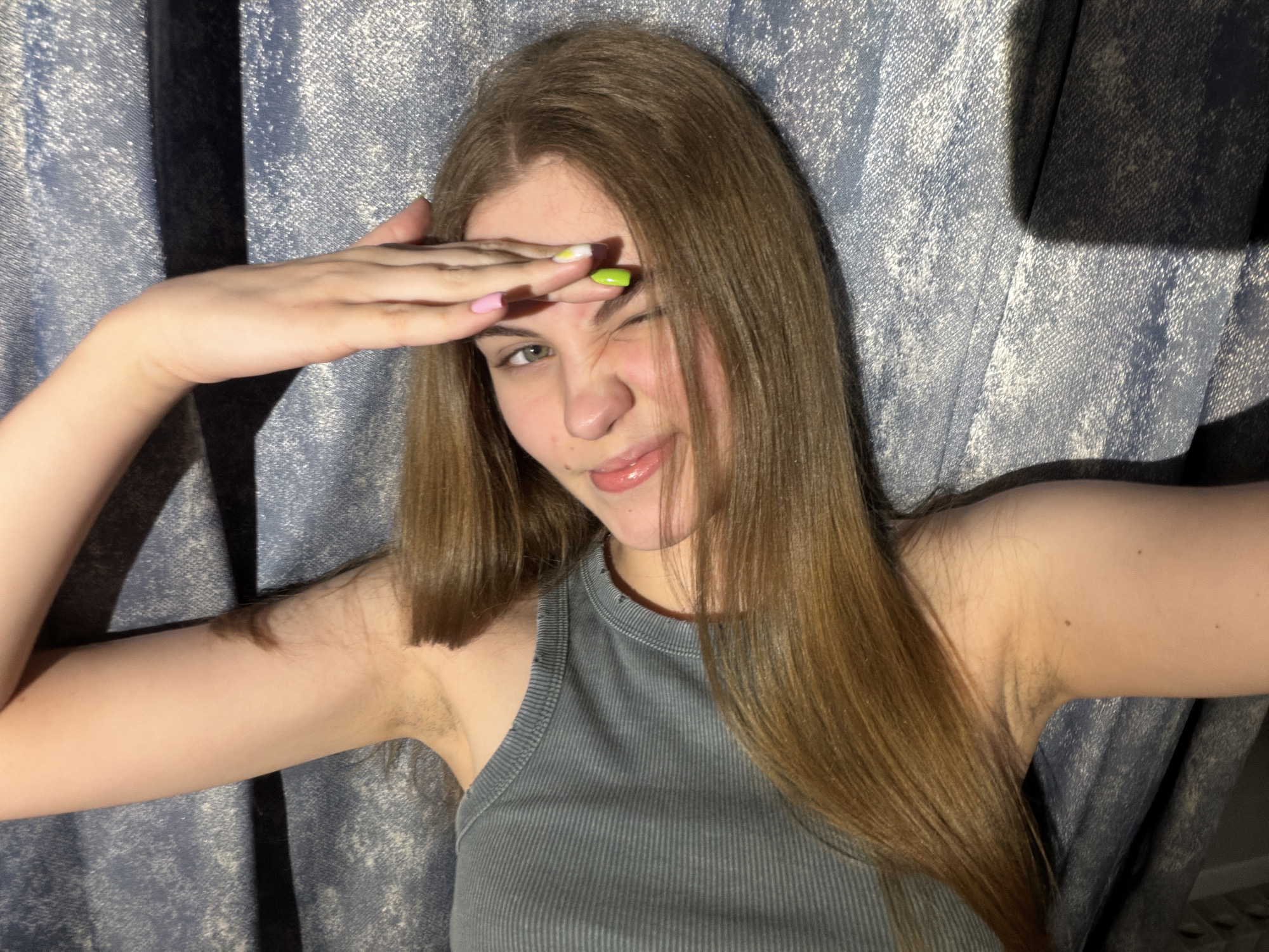 A young woman with long, brown hair and a grey ribbed tank top is shown from the chest up, indoors, against a blue textured backdrop. She is holding her right hand up to her forehead, shielding her eyes. Her left eye is closed in a wink, and her lips are slightly parted, with her tongue visible. The lighting is dramatic, creating shadows on the background and highlighting her facial features. Her fingernails are painted in bright colors. Her left arm is extended outwards, and her right arm is bent, with her hand on her forehead. The background appears to be a curtain or fabric. There is no discernible location in the city of Dnipro, Ukraine.
