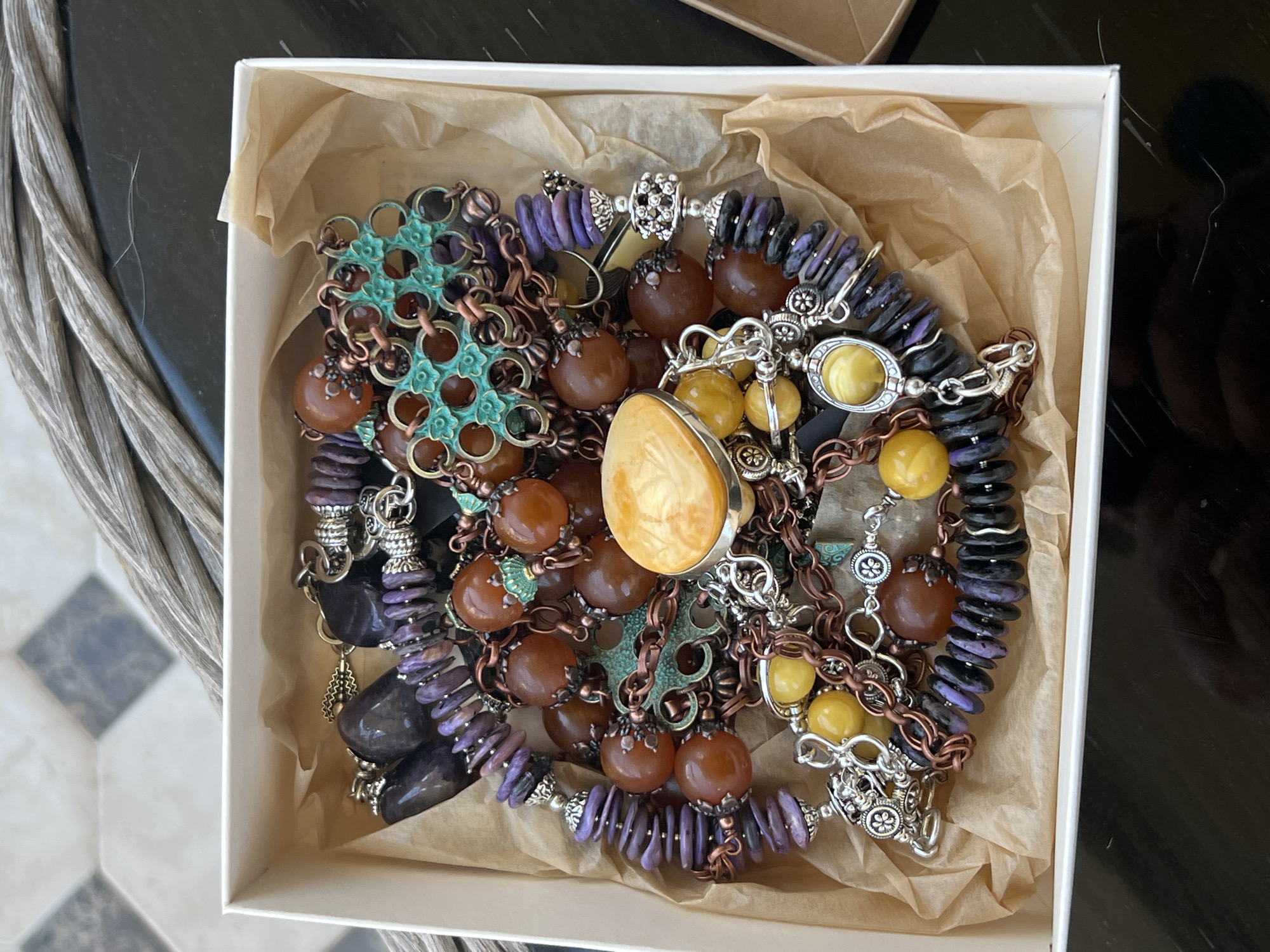 The image shows a close-up, overhead view of a white cardboard box filled with a jumbled collection of necklaces and bracelets. The jewelry features a variety of beads in earthy tones such as amber, brown, and deep purple, interspersed with silver-toned metal components. Some prominent pieces include a large, oval-shaped amber pendant, several strands of round amber-colored beads, and segments of dark purple and black beads. There are also intricate metal findings and chains. The jewelry is resting on crumpled light brown tissue paper within the box. The background of the image includes a dark, reflective surface and a glimpse of a tiled floor with a diamond pattern of grey and white squares. A section of woven material, possibly rope or twine, is visible at the top of the frame. The scene is static, with no people or actions depicted. The overall impression is that of a curated collection of artisanal jewelry. There is no discernible information to locate this scene within the city of Sochi, Russia.