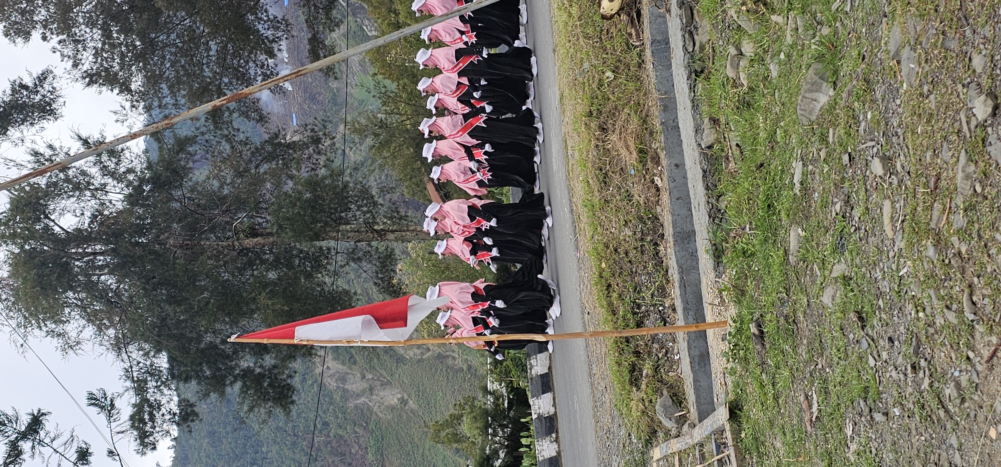 A group of women in black dresses and pink headscarves with red sashes are marching in a procession. They are on a paved road with a grassy embankment on the right. A red and white Indonesian flag is being carried on the left side of the frame. In the background, there are trees and a mountain slope. The scene appears to be in a natural, possibly rural, setting. The attire of the women suggests a formal or ceremonial event, possibly a parade or a cultural gathering. The presence of the Indonesian flag indicates a patriotic or nationalistic context. The mountainous terrain suggests the location is likely in a highland area of Indonesia, potentially Puncak Jaya, known for its mountainous landscape.