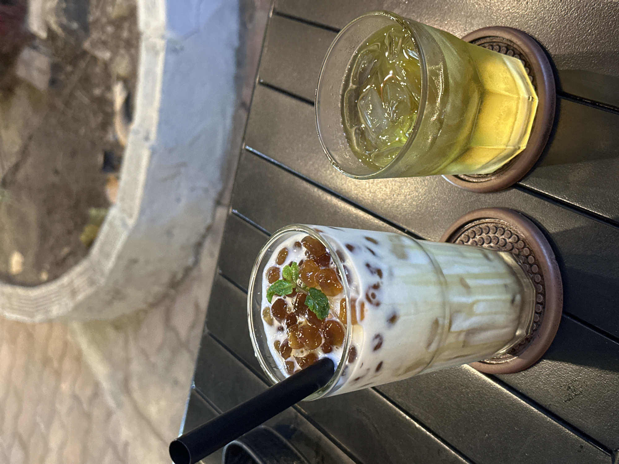 Two refreshing iced drinks are prominently displayed on a dark, grooved table in a casual setting, likely a cafe in Ho Chi Minh City, Vietnam. The scene is captured from a slightly elevated, overhead perspective.
On the top right, a clear glass holds a light yellow-green beverage filled with ice cubes, resting on a round, dark brown coaster with an intricate, textured pattern. The glass shows condensation, indicating its cold temperature.
Below it, towards the center-left, another tall, clear glass contains a milky white drink with numerous dark brown, translucent tapioca pearls settled at the bottom and throughout. A sprig of fresh mint leaves garnishes the top, and a black straw is inserted into the drink. This glass also sits on an identical dark brown coaster.
The table surface is dark, possibly made of wood or metal, with distinct parallel grooves running horizontally. In the soft-focus background, a light-colored, rough-textured structure, possibly concrete or stone, is visible on the left, along with blurred elements suggesting an outdoor or semi-outdoor environment. Natural light suggests it is daytime. There is no visible text in the image.