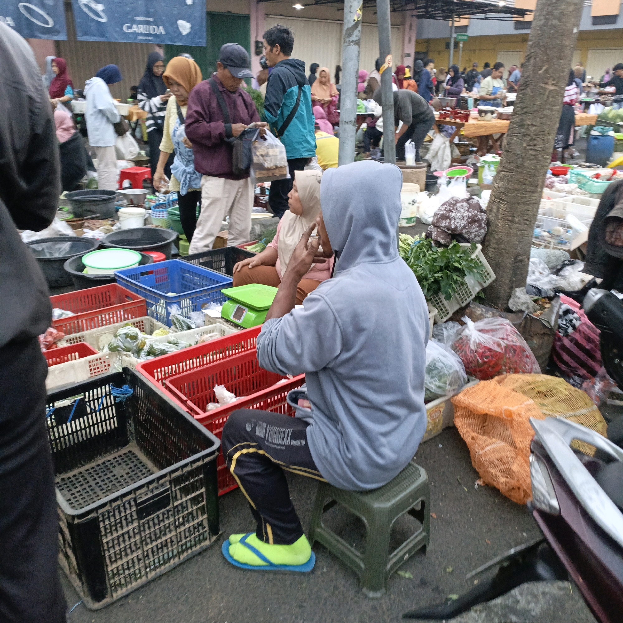 This is a street market scene in Salatiga, Indonesia, with an outdoor setting. In the foreground, a person wearing a grey hoodie and black pants with yellow stripes is sitting on a green plastic stool. They are wearing bright yellow flip-flops with blue straps. In front of them are several plastic crates, some filled with produce like green leafy vegetables and red chilies, contained in plastic bags. A green electronic scale is visible next to the crates.
Behind the seated person, other market-goers are visible, some of whom appear to be vendors. One woman in a yellow hijab and a striped black and white top is looking down, and another person in a purple top is further back. In the background, there are more stalls and vendors, with various goods on display, including baskets of produce and bags of items. The overall impression is a busy marketplace with a variety of people and goods.