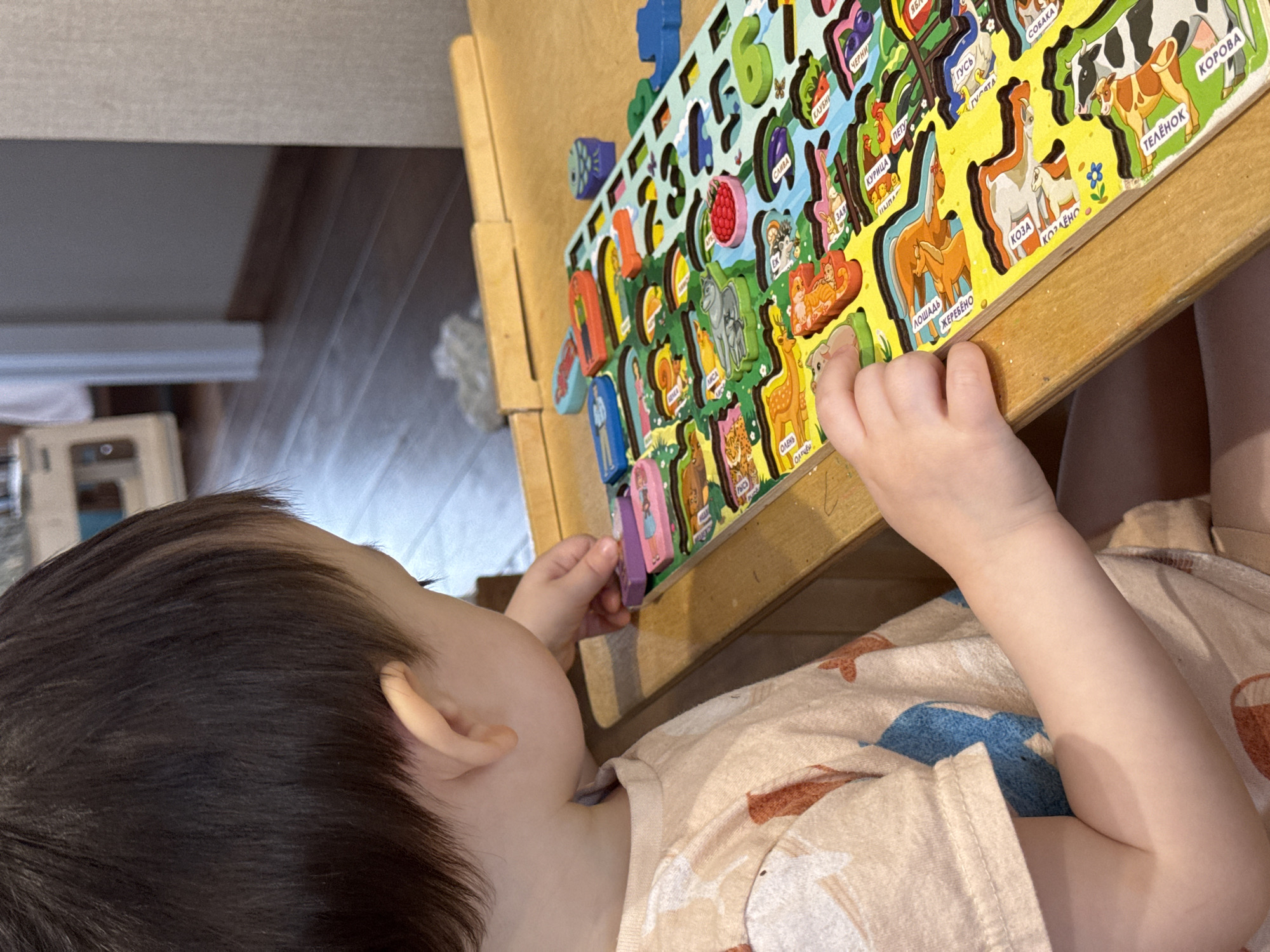 A young child is playing with a colorful wooden puzzle featuring numbers and animals. The puzzle appears to be multilingual, with Russian words visible. The child is fair-skinned and wearing a patterned shirt in light colors. The puzzle is resting on a wooden table. There is no clear information in the image to confirm this location is in the city of Izhevsk, Russia.