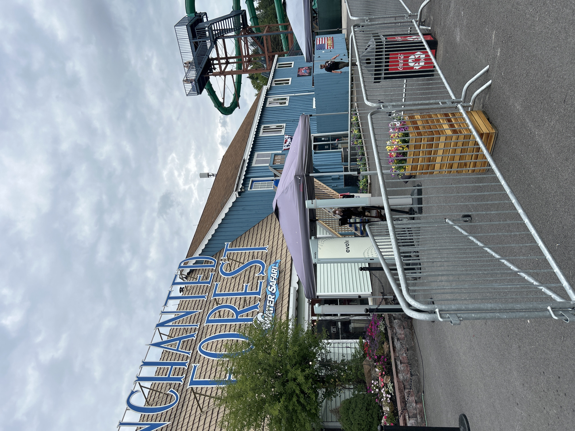 An outdoor scene at Enchanted Forest Water Safari in Old Forge, New York, features a blue building with "ENCHANTED FOREST WATER SAFARI" in large blue letters. A large green water slide structure rises behind the building. In the foreground, metal crowd control barriers line a paved area. A man in a black t-shirt and baseball cap walks past a trash can labeled "Coca-Cola" and a wooden planter filled with colorful flowers. A purple tent stands near the entrance of the building, with another tent visible further to the right. The sky is overcast.