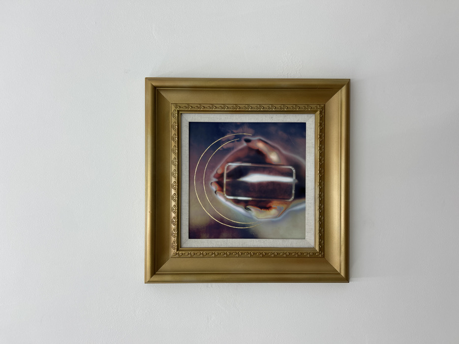 A single square-format artwork, framed in gold, is mounted centrally on a plain, textured white wall. The gold frame features an ornate inner edge with small beaded detailing and a wider, smooth outer profile. Inside the frame, a light beige, fabric-textured mat borders the artwork's central image.
The artwork itself depicts a pair of hands holding a vertically oriented, translucent or reflective rectangular object with rounded corners. The hands, appearing to belong to an adult, show dark nail polish on the fingertips and have a desaturated, slightly reddish-brown skin tone. Three thin, concentric golden rings are superimposed over the upper portion of the hands and the object, emanating from its center. The background of the artwork is a moody, blurred composition of dark blues, purples, and browns, creating an ethereal aura around the central figures. The rectangular object reflects a prominent vertical streak of white light.
The environment is an interior space with diffused, even lighting. The white wall suggests a gallery or museum setting. The specific location within the city of Los Angeles, United States of America, cannot be determined from the visual information provided in the image.