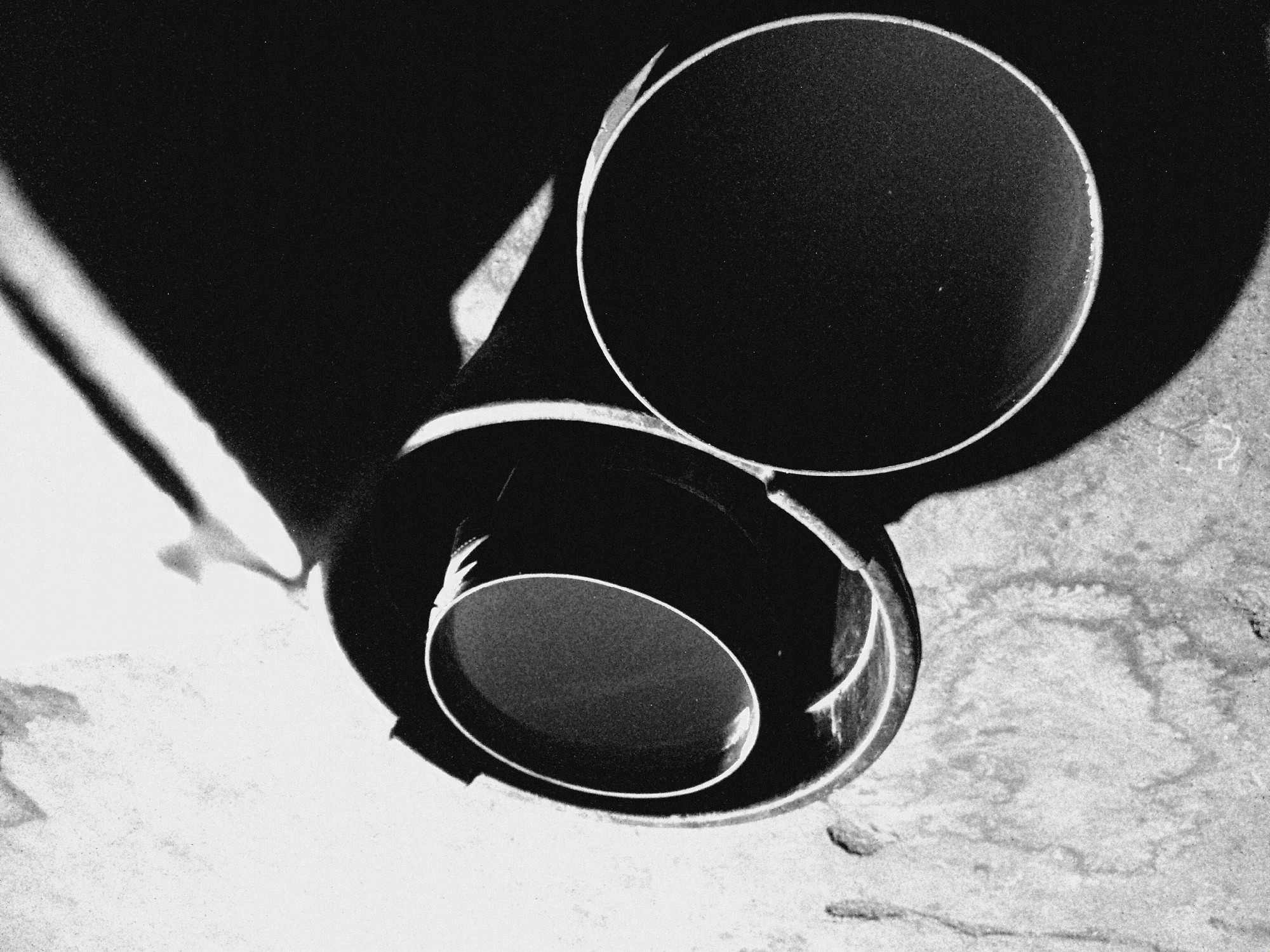 This black and white abstract photograph features two cylindrical objects, likely metal pipes or tubes, arranged on a textured surface. The image is characterized by high contrast and dramatic lighting, creating a play of light and shadow.
A larger cylinder is prominently positioned in the upper right, its circular opening facing upward and slightly left. Its interior appears as a deep, uniform black void, framed by a thin, bright white rim that catches the light. Below and slightly to the left, a smaller cylinder is visible, partially overshadowed. Its interior is also a dark void, with a distinct, luminous oval rim.
Strong light sources create sharp highlights on the edges and surfaces of the cylinders, contrasting sharply with the expansive dark shadows that dominate the background and areas around the objects. A large, geometric shadow stretches across the upper left portion of the frame, emanating from the larger cylinder. The ground beneath the objects is a lighter, textured surface, possibly concrete or stone, displaying a granular quality.
The photograph is taken from an elevated perspective, looking down onto the arrangement. The composition emphasizes geometric forms – circles, ovals, and linear shadows – and the stark tonal differences between light and dark. There is no discernible activity taking place, and no people or visible text are present. The time of day, weather, or specific location cannot be determined from the image.