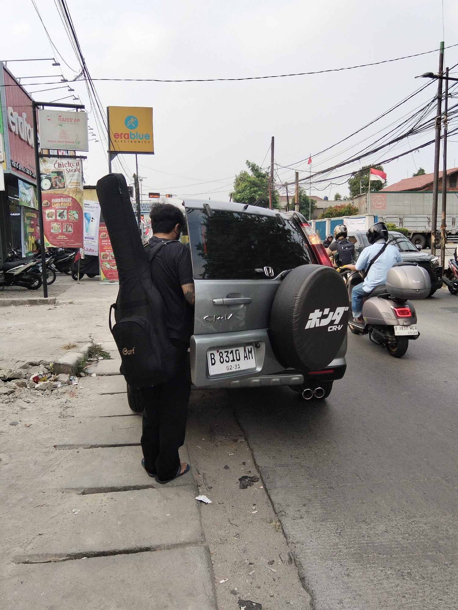A man stands on a sidewalk next to a silver Honda CR-V, holding a black guitar case. The man is wearing a black t-shirt and black pants, with a tattoo visible on his left arm. The guitar case has the brand "Cort" printed on it. The CR-V has a spare tire cover with Japanese lettering. In the background, a grey scooter with a rider wearing a blue shirt and jeans is moving on the road. There are other vehicles and buildings visible, including a truck. The scene appears to be on a street in South Tangerang, Indonesia, as indicated by the license plate of the CR-V which reads "B 8310 AM". The environment includes a sidewalk with cracked concrete, debris, and power lines overhead. A yellow billboard with the "erablue ELECTRONICS" logo is visible in the distance.