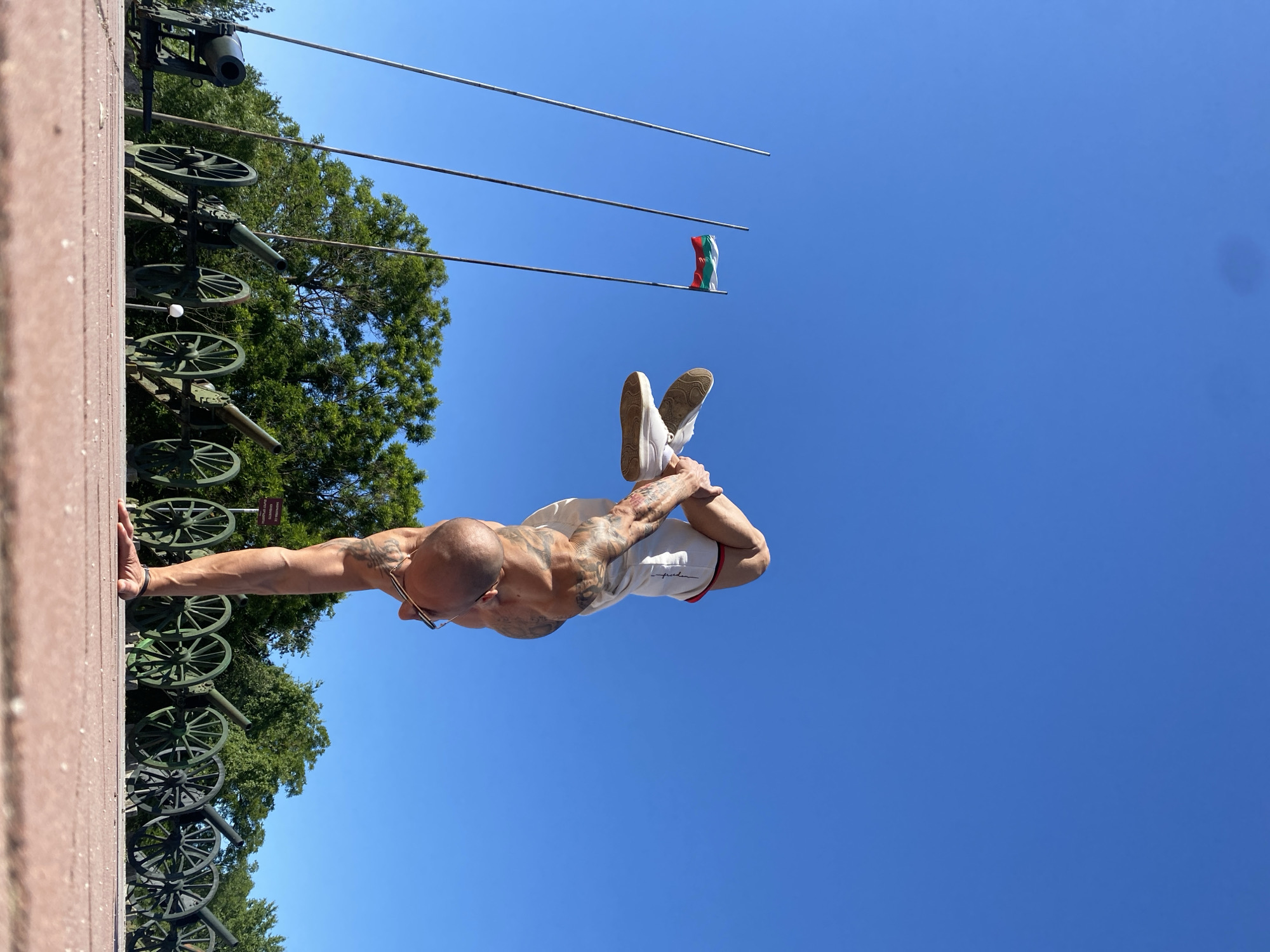 A man performs a handstand on a brick wall in front of a cannon display at the Pleven Epopee 1877 Memorial Park in Pleven, Bulgaria. The man has a bald head, glasses, and tattoos on his torso and arms, and he wears white shorts and shoes. A Bulgarian flag is visible on a pole in the background. The sky is blue, and there are trees nearby.