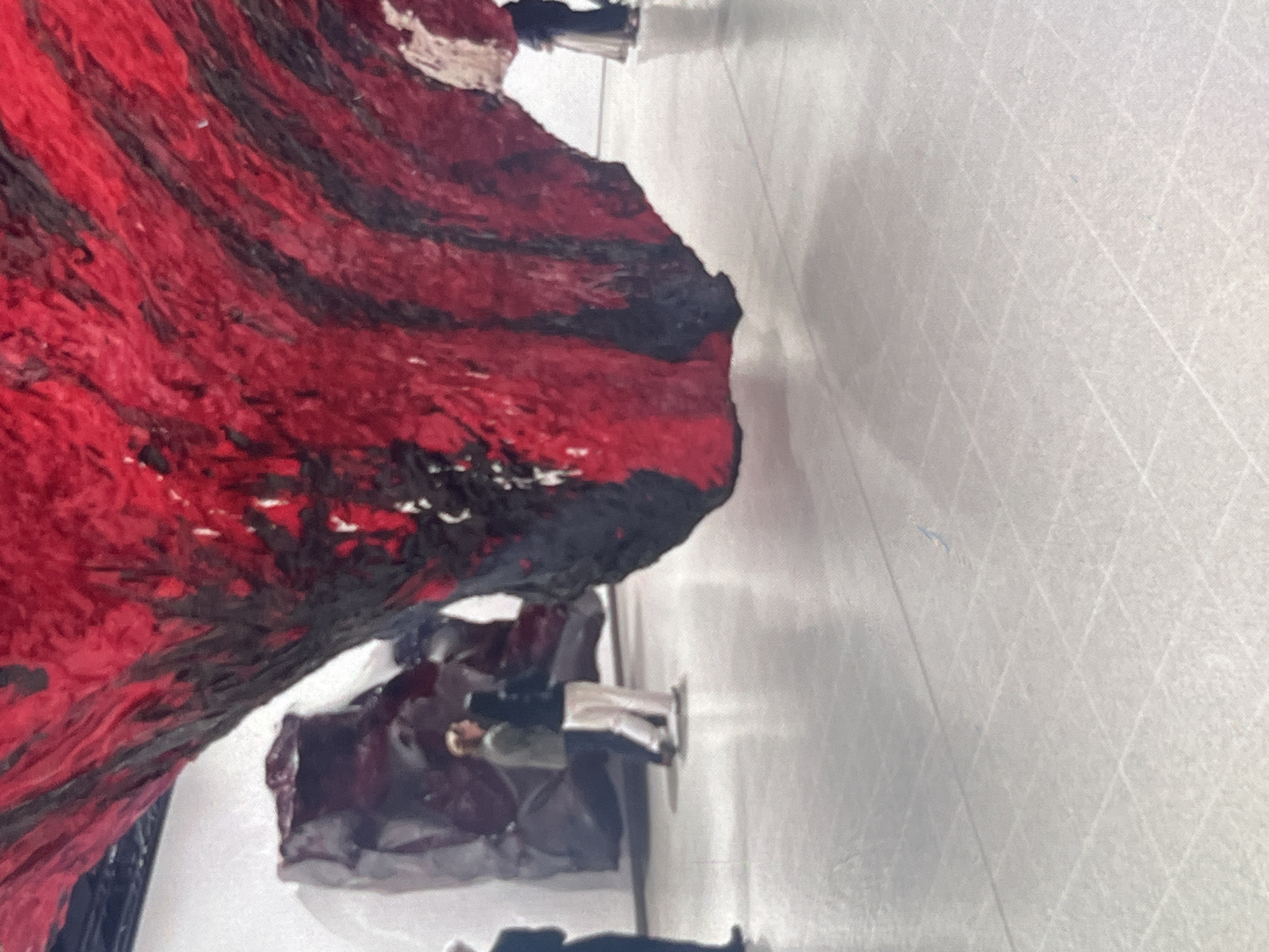 The image depicts an indoor exhibition space, possibly an art gallery, with a large, abstract sculpture dominating the foreground. The sculpture is characterized by its textured surface and bold red and black coloration, suggesting organic or raw materials. In the background, two individuals are observing the artwork. One is wearing a green jacket and light-colored pants, while the other is in a dark hoodie and light pants. Further in the background, another figure in a dark dress stands near a dark, boulder-like structure. The floor is a light-colored, polished surface marked with a grid pattern. The scene is illuminated by artificial lighting, casting soft shadows. The location in Stepantsminda, Georgia is not evident from the visual information provided in the image.
