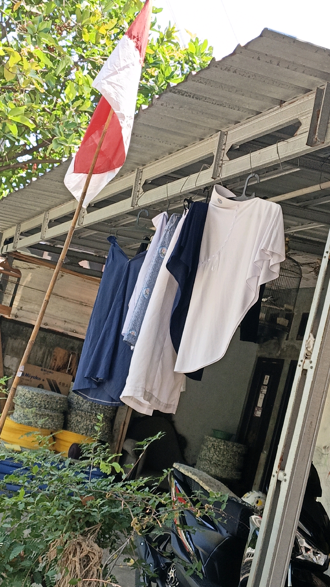 A low-angle, slightly tilted outdoor shot captures an Indonesian flag flying from a bamboo pole, leaning against a corrugated metal roof structure. Several articles of clothing hang from hangers on a metal rail beneath the roof. These include a dark blue dress, a white patterned top, and a light pink asymmetrical tunic worn over a dark garment. A birdcage is visible to the right of the clothing. In the foreground, lush green foliage obscures a black motorcycle. The background reveals stacked circular objects, some yellow and others gray and speckled, and a wooden structure. The overall impression is one of a somewhat cluttered, lived-in outdoor space, possibly a backyard or alley in a residential area of Cikal, Indonesia.