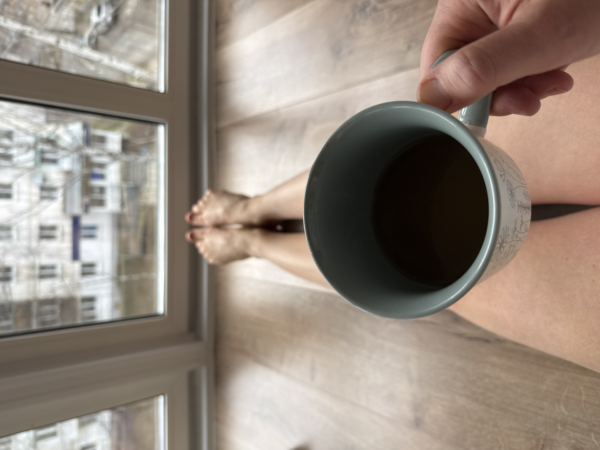A first-person perspective image shows a person seated on a light brown wooden floor with legs outstretched towards a window. The feet are visible with red painted toenails. A blue-grey patterned mug, held by a hand, is in the foreground. The mug contains a dark liquid, likely coffee or tea. The window reveals an exterior scene with buildings visible in the reflection, suggesting an urban environment. Based on the architecture, specifically Soviet-era housing blocks visible through the window and the general appearance, the location is likely Chaikovsky, Russia. The weather appears overcast.