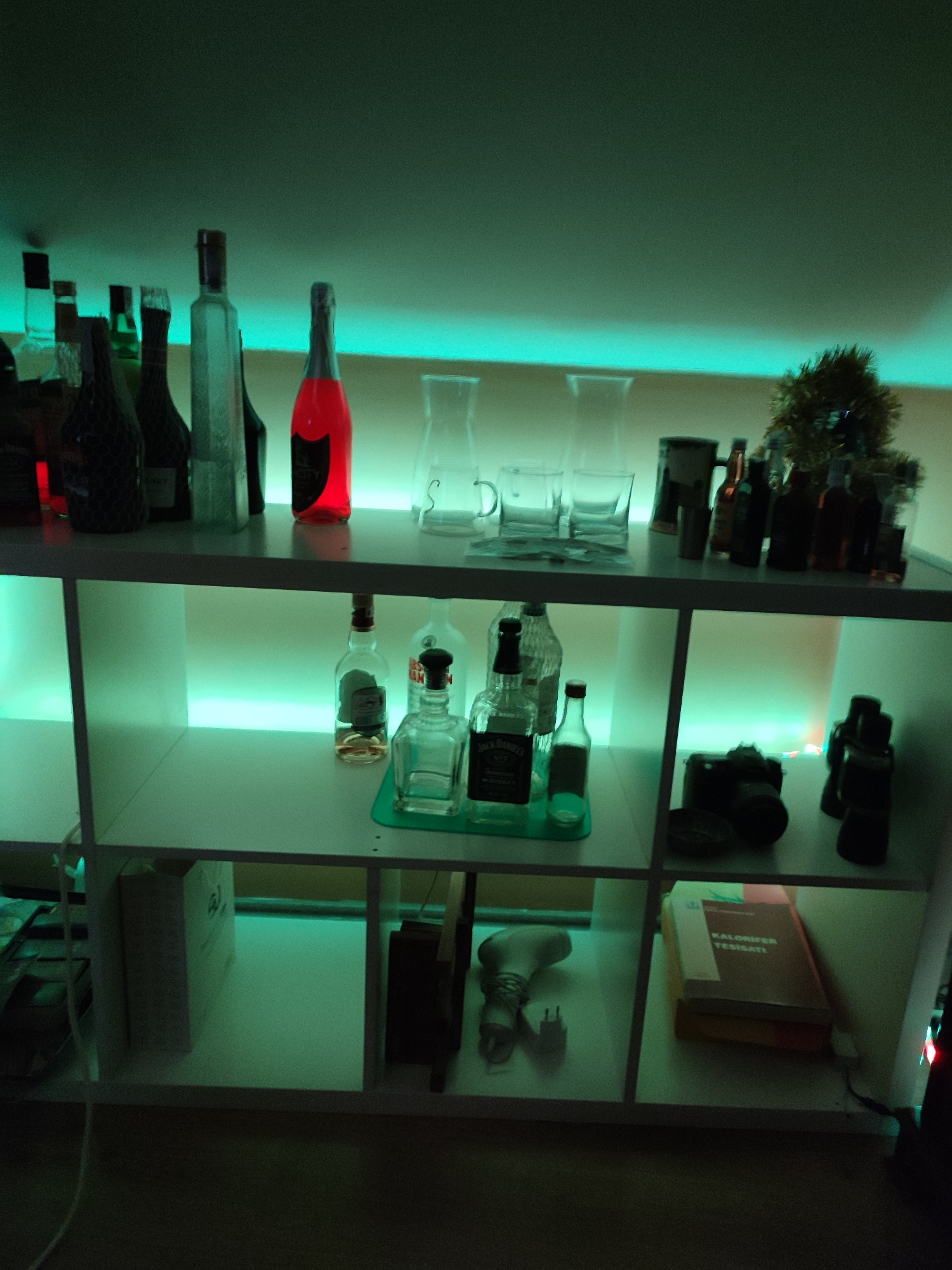 The image depicts an indoor scene, likely a home or bar, with shelves illuminated by green and cyan lighting. The shelves are stocked with various items, primarily alcohol bottles, glasses, and some decorative objects.
On the top shelf, there are several bottles of alcohol, including a prominent red bottle, and various glasses. A small, glittery tree-like decoration is also visible.
The middle shelf holds more bottles and glasses. Notably, there are several clear glass bottles, some containing amber-colored liquid, and a clear square bottle.
The bottom shelf contains a few larger items. In one section, there is a black camera. In another, there is a book or box with text that appears to be in Turkish. A light bulb and plug are also present in one of the compartments.
The overall environment appears to be dimly lit, with the colored lighting creating a mood. There are no people visible in the image. The furniture is white and modular, resembling a cube shelf unit. The background wall is out of focus and illuminated by the colored lights. The image is shot from a slightly elevated angle, looking down onto the shelves.
