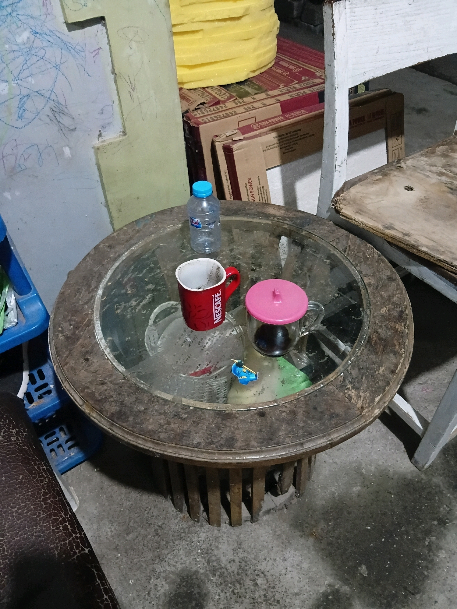 In Cikal, Indonesia, a circular wooden table with a glass top sits on a concrete floor. On the table are a small clear glass mug with a pink lid containing a dark liquid, a red Nescafe mug, and a clear plastic bottle of water. The table is positioned near a wall with scribbled drawings. A wooden chair with a worn seat is partially visible to the right, and to the left, a blue plastic shelf unit stands. Cardboard boxes and a stack of yellow material are visible in the background. The scene appears to be an indoor or covered outdoor space.